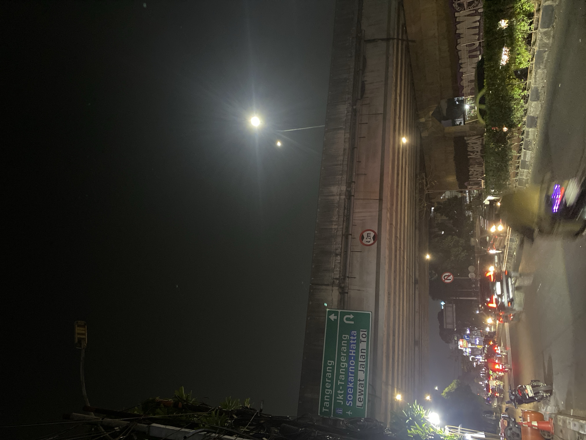 The image captures a street scene at night in Tangerang, Indonesia, under an elevated road or bridge. A green traffic sign indicates directions: "Tangerang" at the top, followed by "Jkt-Tangerang" with an icon of a building, and "Soekarno-Hatta" with a plus sign, and "Lewat Jalan Tol" with an arrow pointing to the right. To the right of the sign, a circular red sign displays "4,2m", indicating a height restriction. The road below is filled with traffic, including numerous motorcycles and cars. Streetlights illuminate the scene, casting a glow on the road and the understructure of the bridge. The environment appears to be an urban setting with buildings and greenery visible in the background. The overall impression is a busy, nighttime cityscape.