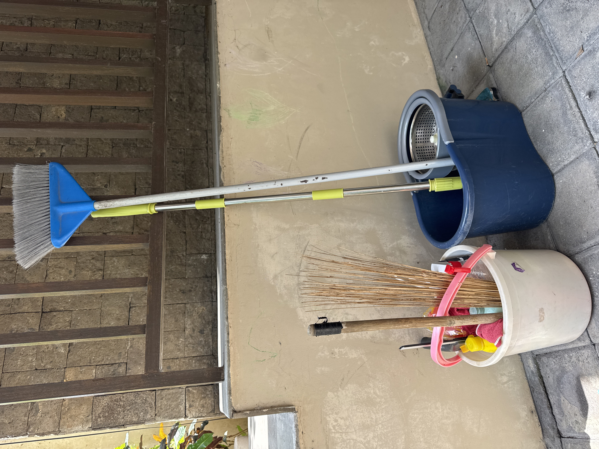 The image depicts a collection of cleaning supplies in an outdoor area. A blue mop bucket with a spin-drying mechanism is positioned next to a white bucket containing a variety of items, including a straw broom and cleaning cloths. A push broom leans against a wall. The setting is a concrete or paved area adjacent to a building, possibly in the city of Jimbaran, Indonesia, based on the typical architectural style and materials found in the region. The scene appears to be a storage area for cleaning tools. No people are visible.