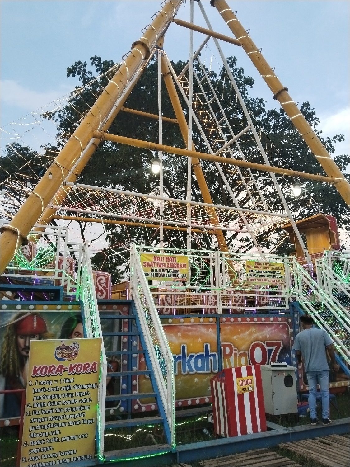 This evening scene in Matesih, Indonesia, captures a vibrant amusement park ride, likely a Kora-Kora (pirate ship) pendulum ride, illuminated against a fading sky. The large, yellow metal structure of the ride dominates the frame, adorned with numerous string lights that cast a warm glow. Behind it, dark green tree foliage is visible against the light blue and grey sky, indicating dusk.
A man in a blue t-shirt and jeans stands on a wooden platform near the ride's entrance, facing away from the viewer. Steps with white handrails lead up to the loading area of the ride, which is decorated with colorful panels. One panel features an image resembling Captain Jack Sparrow, and another part of the ride's branding displays "berkah Ria 07".
Several signs provide information and warnings. A prominent yellow sign on the left lists rules for the "KORA-KORA" ride in Indonesian:
1. 1 person = 1 ticket (children must be accompanied, companions must still pay).
2. Must be in good health.
3. Pregnant women and visitors with heart disease/high blood pressure are not allowed to ride.
4. Sit orderly, hold tight.
5. Watch your belongings (Loss is not the responsibility...).
Another banner above the stairs reads, "HATI HATI SAAT MELANGKAH JATUH DI TANGGA AK SEINDAH JATUH CINTA" (Be careful when stepping, falling on stairs is not as beautiful as falling in love). Adjacent text praises the ride, mentioning its excitement and thanking visitors. A red and white striped box displaying "10.000" (likely a price or denomination) stands near the entrance. The scene depicts a lively, local carnival atmosphere at the close of day.