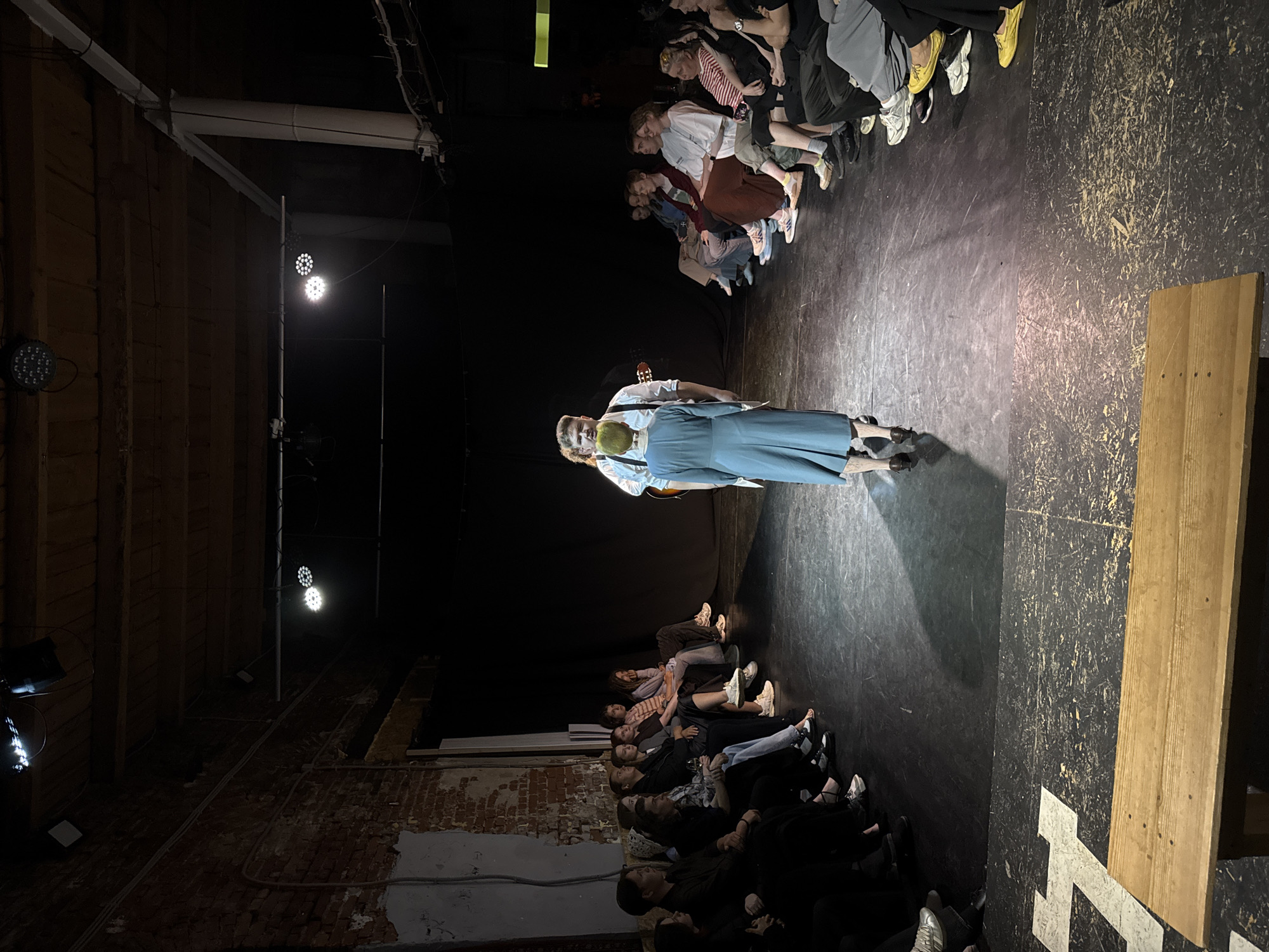 A dimly lit performance space appears to be set in Izhevsk, Russia. The scene is viewed from an elevated perspective, likely from the rear of the stage. A figure in a light-colored shirt and blue overalls is standing center-stage with a guitar. Audience members line either side of the performance space, seated on risers or benches, facing the performer. Exposed brick and wooden beams are visible on the sides of the space, suggestive of a raw, possibly repurposed industrial building. Stage lighting illuminates the central area where the musician is standing.