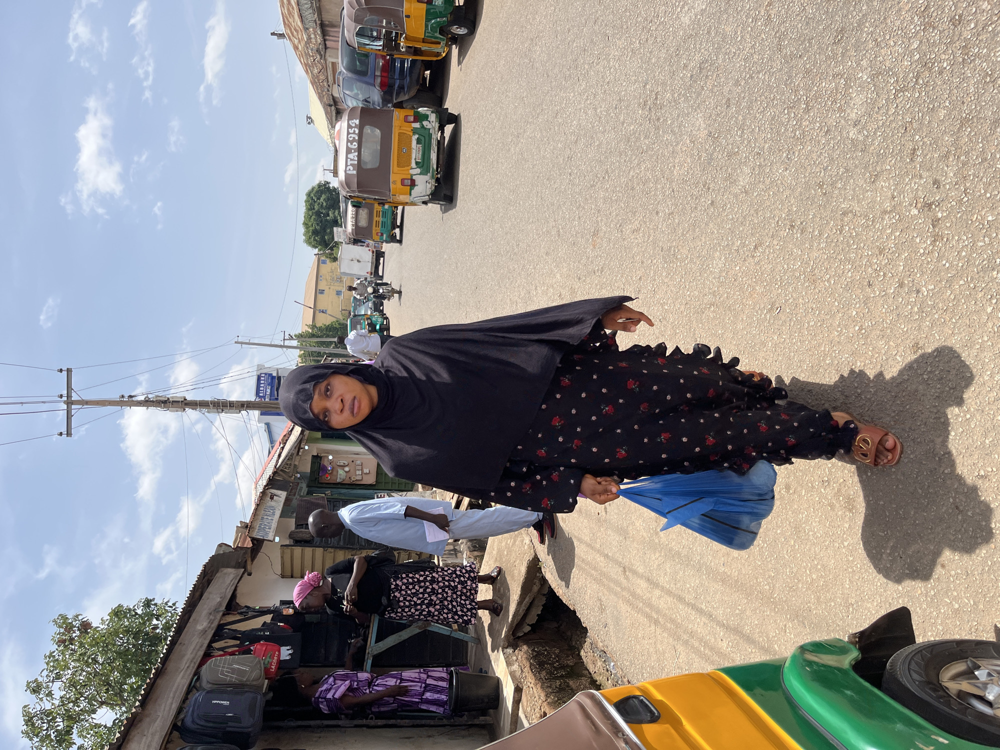 A woman in a black hijab and a long black dress with red floral patterns walks down a street in Jos, Nigeria. She is holding a blue plastic bag. Shops line the left side of the street, displaying luggage and other goods. Several people are standing or walking in front of the shops. Vehicles, including auto-rickshaws (tuk-tuks), are parked along the street. Overhead power lines are visible against a partly cloudy sky. The pavement is cracked and shows signs of wear.