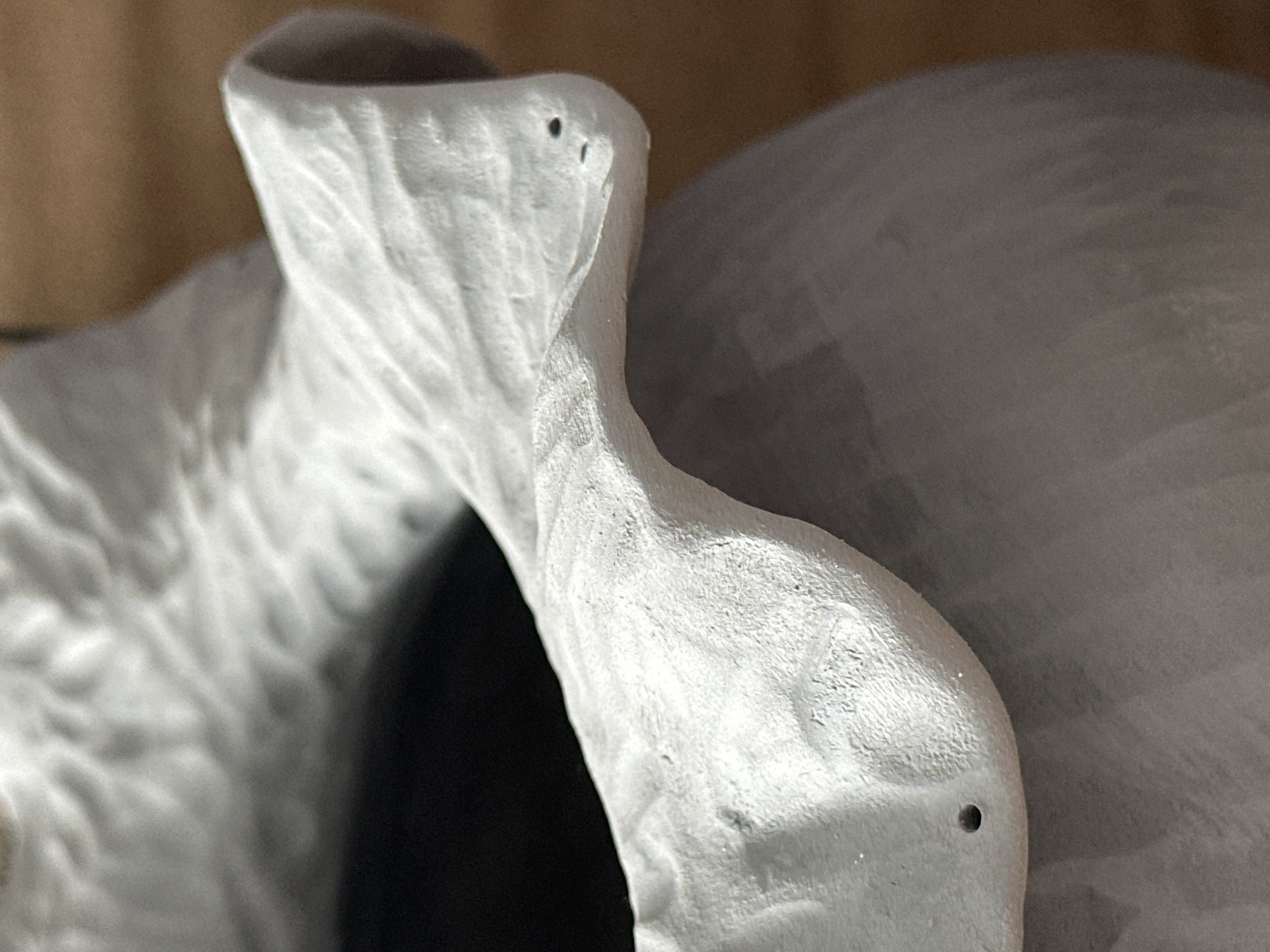 The image is a close-up, macro photograph of a textured, grey ceramic vessel. The vessel has a ruffled or petal-like rim with visible indentations and small holes, suggesting it might be a decorative vase or pot. The texture is rough and uneven, with some areas appearing smoother than others. The interior of the vessel is dark and out of focus. The background is a blurred warm brown color, indicative of a wooden surface. There are no people or specific landmarks visible that would allow for localization to Nevinnomyssk, Russia or any other specific location. The lighting appears to be natural, coming from the side, creating subtle shadows and highlighting the texture of the ceramic.