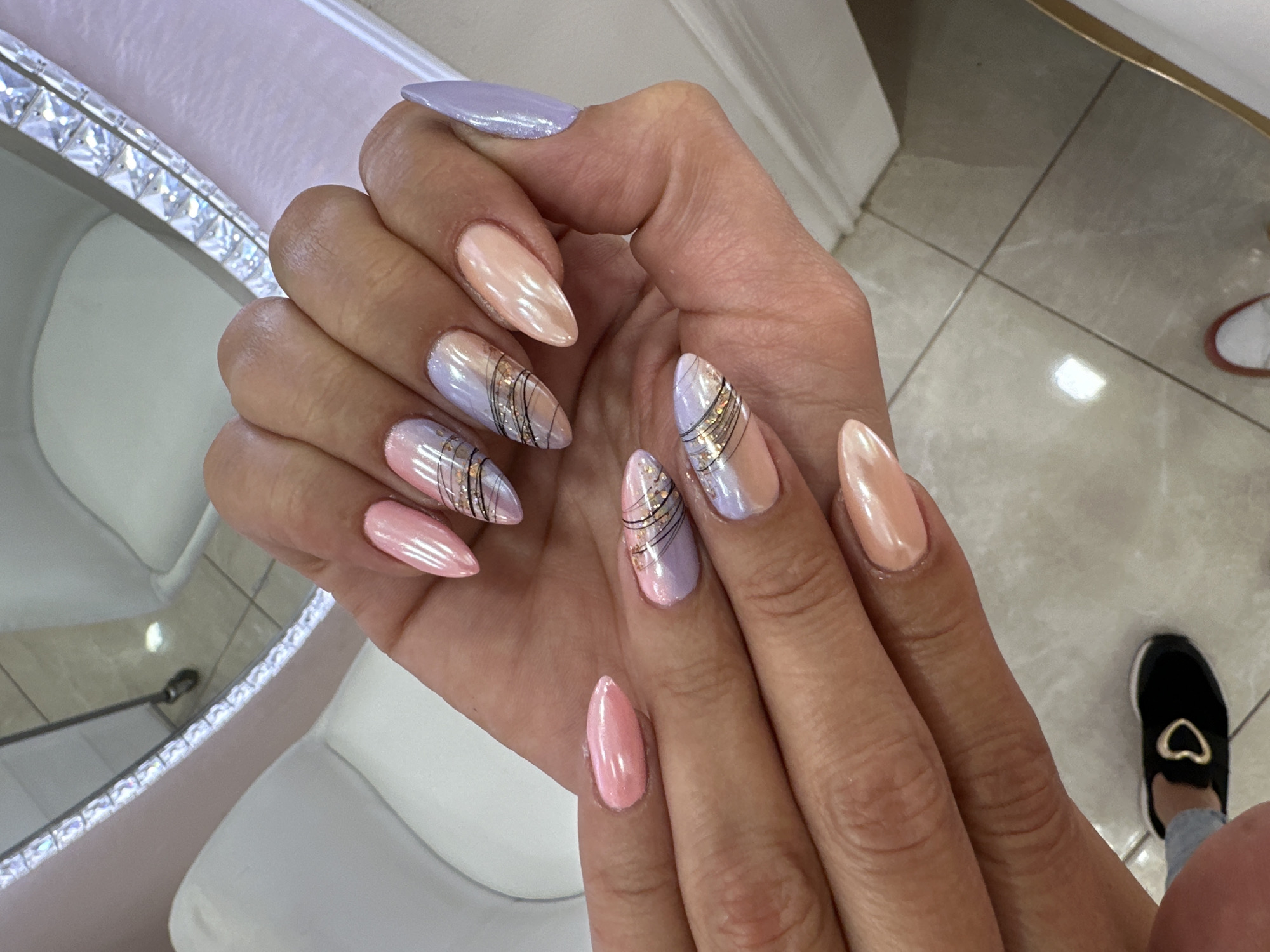 The image depicts a close-up view of a person's hand with manicured nails. The nails are almond-shaped and painted in various pastel colors (lilac, peach, pink). Some nails feature an intricate design of thin gold and black lines. The person is in an indoor setting with tile flooring visible in the background. A chair with a reflective edge is also visible. A black slip-on shoe with a gold heart-shaped adornment is partially visible in the lower right corner of the image. It's impossible to verify the location is Sofia, Bulgaria from visual information alone.