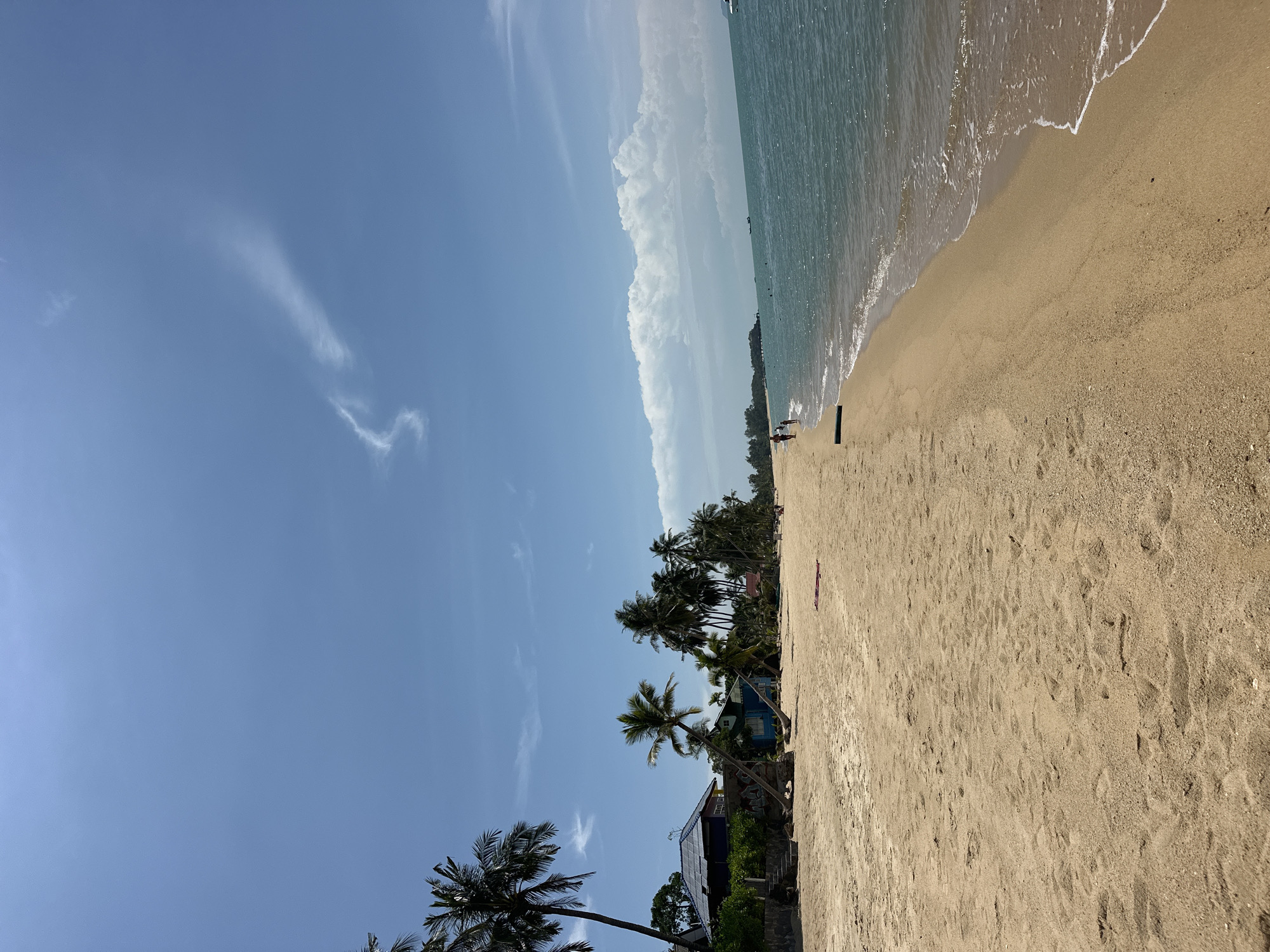 A wide shot captures a sandy beach on a bright, sunny day in the Ko Samui District of Thailand. The sand, appearing a light tan, stretches across the foreground and mid-ground. Several palm trees, with their distinctive fronds, are scattered along the edge of the beach, some leaning towards the water. Behind the trees, a row of small, colorful houses can be seen, painted in blues and purples, with some appearing to be bungalows or beach huts.
The ocean occupies the right side of the frame, its water a vibrant turquoise color. Gentle waves with white foamy crests lap at the shore. In the distance, a few boats are visible on the horizon, along with a line of hazy landmass. The sky is a clear blue, with a few wispy white clouds present, one particularly elongated cloud stretching across the upper portion of the image.
A few people are present on the beach. Two individuals are walking along the water's edge in the mid-ground on the left side of the frame. Another person appears to be walking on the sand further down the beach to the right. A small, rectangular object, possibly a mat or towel, is lying on the sand in the mid-ground. The scene conveys a relaxed, tropical atmosphere.