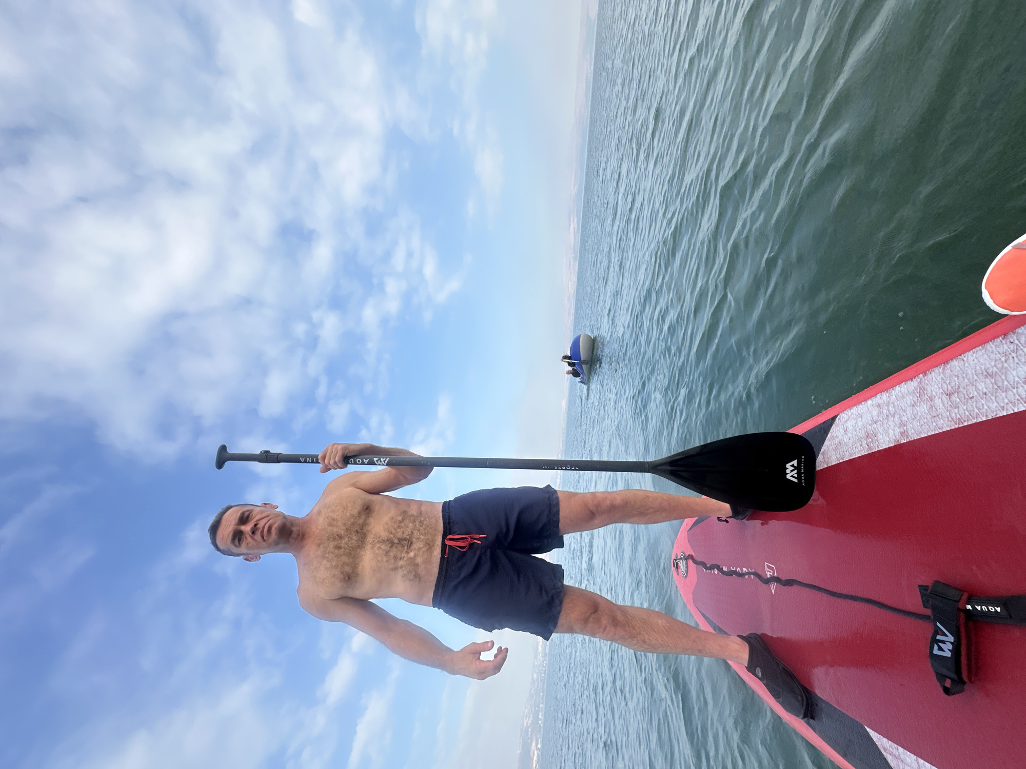 A shirtless man stands on a red paddleboard in the Sea of Galilee, near the city of Tiberias, Israel. He is holding a paddle with a black blade and a logo. The man is wearing dark blue swim shorts with a red drawstring and black water shoes. In the background, another small boat with two people is visible on the water. The sky is blue with scattered clouds. The overall scene depicts a recreational activity on the water.