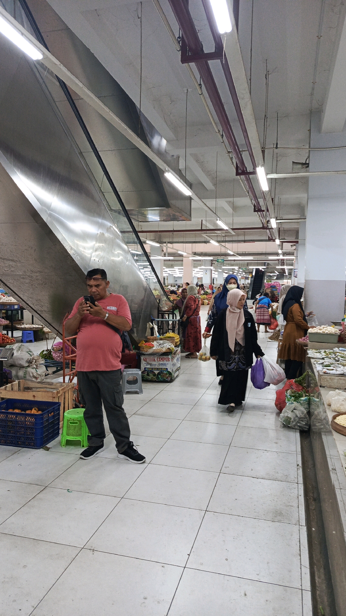 A man in a red t-shirt and dark pants is standing near an escalator, looking at his phone. He is surrounded by market stalls and other shoppers. The market appears to be indoors, with a tiled floor and fluorescent lighting. In the background, there are more stalls selling produce and other goods. The scene is busy and active, with people moving about and interacting with each other. The location is specified as Nologaten, Indonesia.