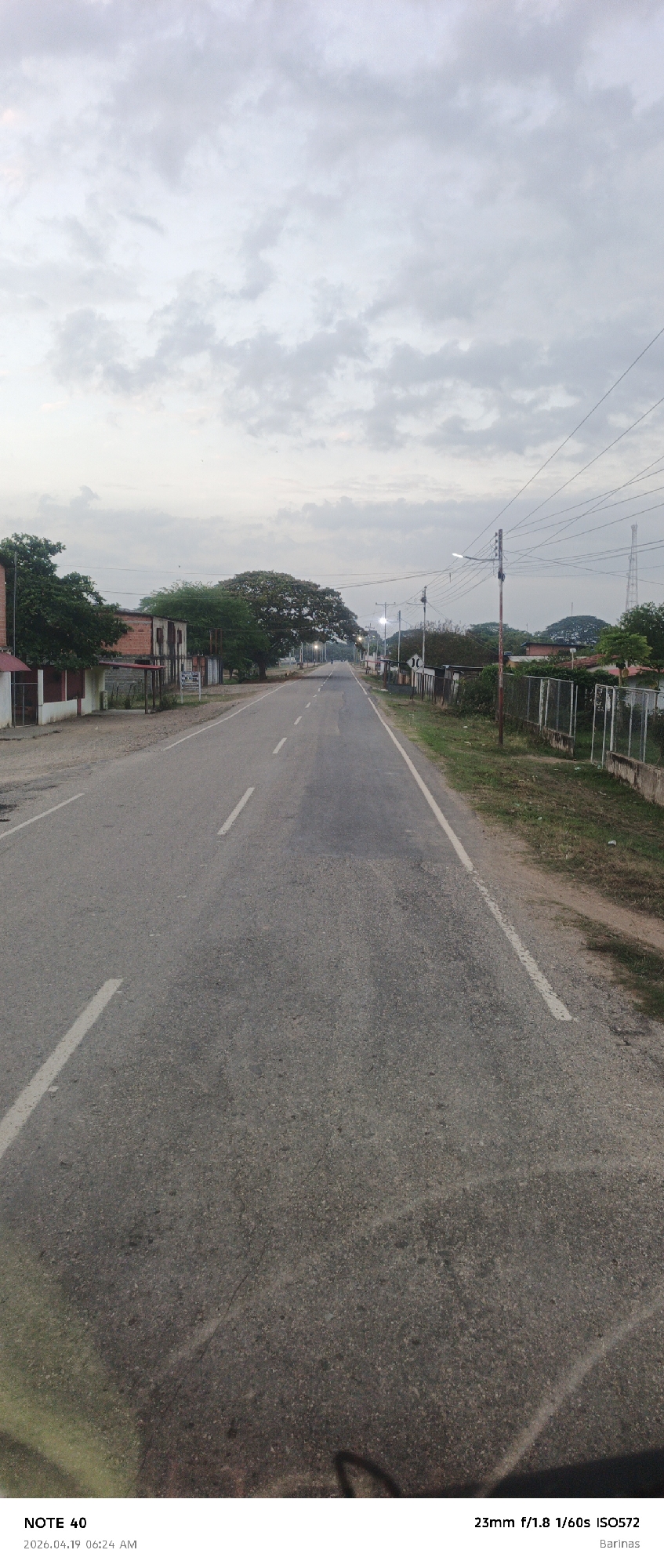 The image depicts a straight asphalt road stretching into the distance under a cloudy sky. The road has two lanes with dashed white lines separating them. The pavement shows signs of wear and repair. On either side of the road, there are grassy areas and low buildings, some appearing residential. Utility poles and wires run along the roadside. A tall communication tower is visible in the distance. The metadata indicates the image was taken with a Note 40 camera, using a 23mm lens with an aperture of f/1.8, a shutter speed of 1/60s, and an ISO of 572, in Barinas, Venezuela. Based on the visual information, it is not possible to determine the exact location within the city of Arismendi, Venezuela. There are no people visible in the image, and the scene appears static.