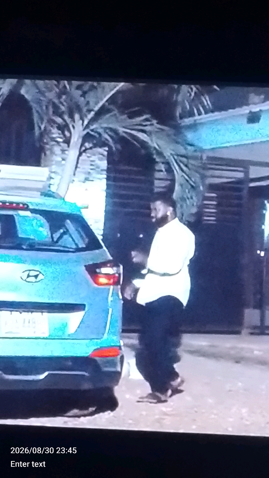 In Lokoja, Nigeria, at 23:45 on August 30, 2026, a man stands beside a light blue car. He is wearing a white long-sleeved shirt and dark trousers. He is facing to his right, away from the car, and appears to be in the process of exiting the vehicle. The background shows a palm tree and a building with horizontal slatted elements, suggesting an outdoor, possibly residential or commercial, setting in a tropical or subtropical urban environment. The overall lighting suggests it is nighttime.