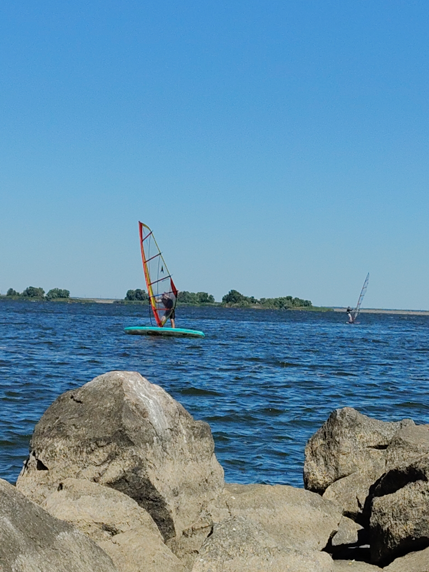 The image depicts a serene day on a body of water, likely a river or lake, with two individuals engaged in windsurfing. In the foreground, large, weathered rocks form a natural barrier along the shore. The water is a deep blue, reflecting the clear, cloudless sky above.
The primary subject is a windsurfer positioned in the middle ground, actively steering their board with a colorful sail that catches the sunlight. The sail exhibits a combination of red, yellow, green, and blue panels, with a dark silhouette of the windsurfer visible behind it. The windsurfer appears to be wearing dark clothing.
Further in the background, a second, smaller windsurfer is visible, positioned to the right of the first. This windsurfer's sail is primarily white and gray, and they are also actively engaged in the sport.
The distant horizon is marked by a strip of land covered in lush green trees, suggesting a natural or park-like setting. The overall atmosphere is one of peaceful outdoor recreation. While the exact location within Cherkasy, Ukraine, cannot be definitively determined from the image alone, it strongly suggests a waterfront area suitable for water sports, such as the Kremenchuk Reservoir, which is adjacent to the city and known for such activities.