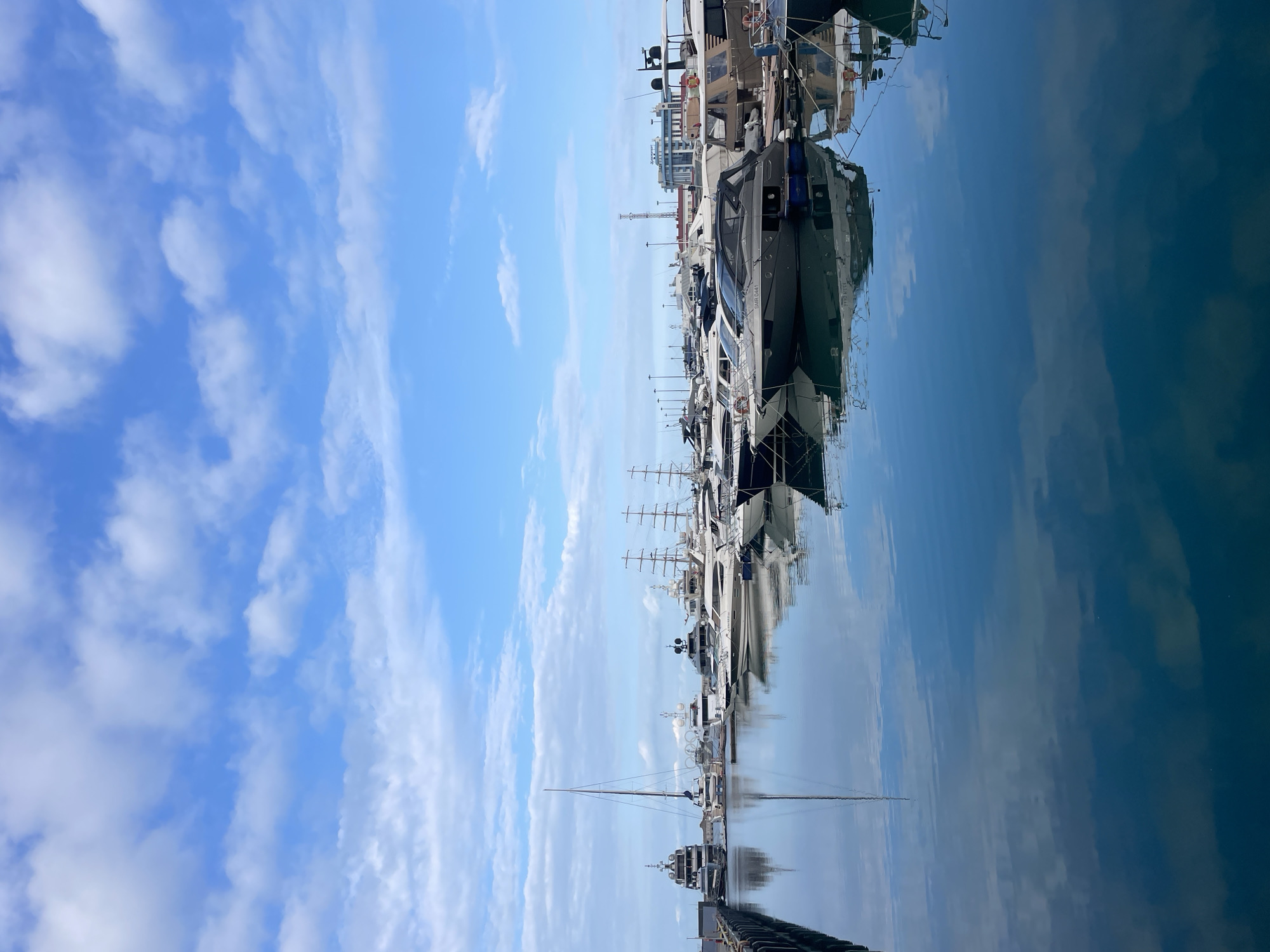 The image depicts a marina in Sochi, Russia, filled with various yachts and boats docked in calm, reflective water under a blue sky with scattered white clouds. In the foreground, a dark wooden pier juts into the frame from the left. Behind it, a fleet of vessels, ranging from sleek modern yachts to larger sailing ships, are neatly arranged. Several of these yachts have distinct, modern designs with dark hulls and polished surfaces, catching the light. Tall masts of sailing yachts are prominent features in the mid-ground. In the background, several buildings, including what appears to be a hotel or administrative building with a blue roof, and some telecommunication towers, are visible along the shoreline, suggesting an urban setting. The water mirrors the sky and the boats, creating a serene and picturesque scene. No people are visible in the image.