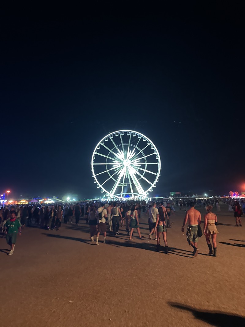 The image captures a nighttime scene at the Coachella Valley Music and Arts Festival in Indio, California, United States. A brightly lit Ferris wheel dominates the background. In the foreground, a large crowd of people is dispersed across a flat, dirt surface. The people are dressed in casual festival attire, and their shadows are elongated by the low lighting. The crowd thins out towards the back. There are lights and structures of other festival attractions visible behind the crowd. The sky is dark and clear.