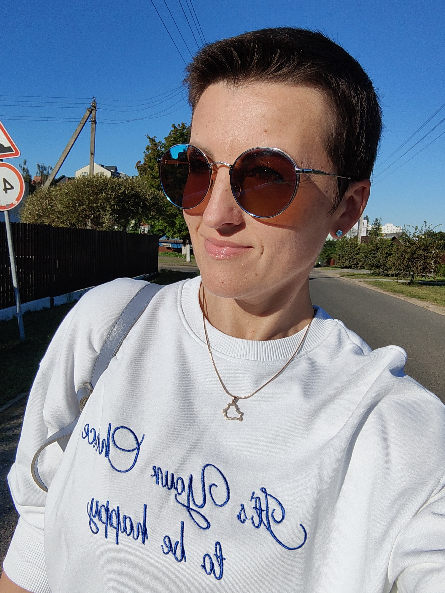 In an outdoor, daylight scene in Fanipal, Belarus, a woman with short, dark hair and sunglasses is captured in a selfie. She is wearing a white sweatshirt with blue embroidered text that appears to be a quote. A delicate necklace with a star-shaped pendant is visible. The background shows a paved road, a wooden fence, trees, houses, and a utility pole with power lines against a clear blue sky. A traffic sign with a "4" in a circle is also present on the left side of the frame. The woman is holding a light gray bag over her shoulder.