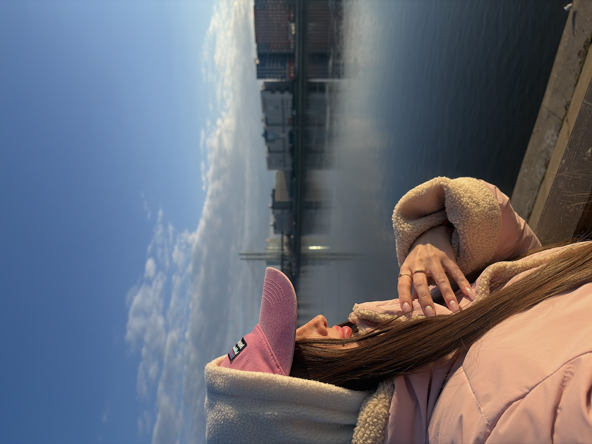 A young woman is lying on a concrete structure, with a cityscape in the background, appearing to be in Saint Petersburg, Russia. The woman is wearing a pink jacket with a sherpa lining and a pink baseball cap. Her long, dark hair is spread across the jacket. She is wearing a gold ring on her right hand. In the background, there is a body of water reflecting a cityscape under a blue sky with clouds. The photo is taken from a high-angle perspective.