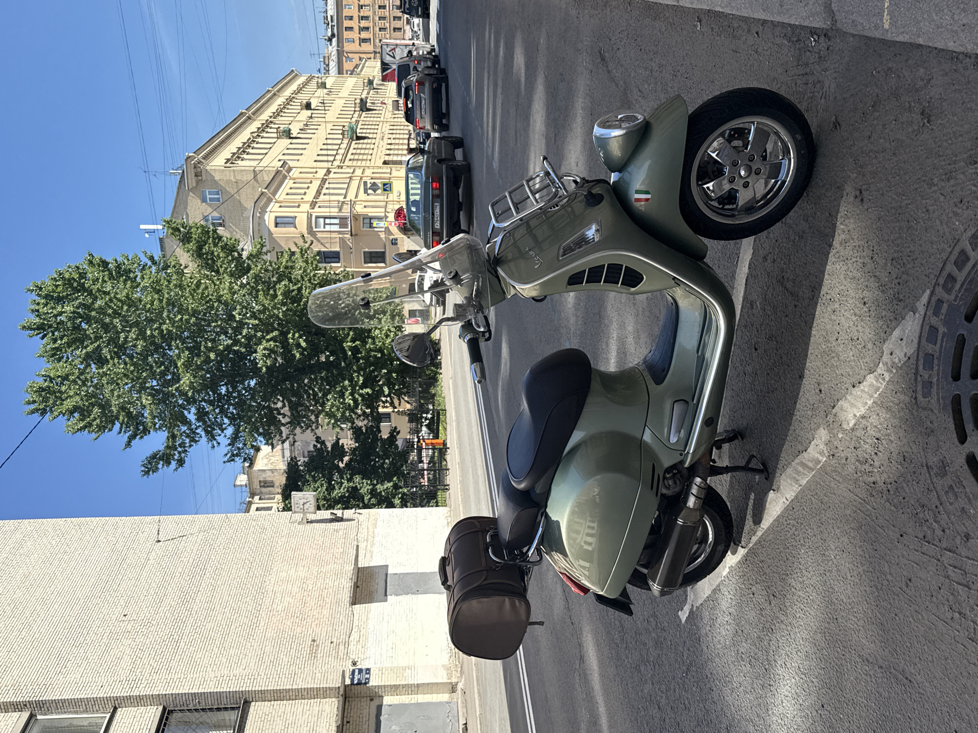 The image displays an urban street scene bathed in bright daylight under a clear blue sky. The main subject is a light olive green Vespa scooter, parked on the side of an asphalt road, partially on a concrete sidewalk. The scooter is equipped with a clear windshield, a chrome front luggage rack, a black seat, and a brown cylindrical storage bag attached to its rear. Notable details on the scooter include its chrome wheels, a "Vespa" logo on the front, and a small Italian flag decal on the front fender.
In the background, the street extends, lined by multi-story buildings. One prominent building on the right has an ornate, light-colored facade with many windows, showcasing a classic architectural style. On the left, another building features a light-colored, textured brick exterior. A large, leafy green tree stands prominently on the left, offering a natural element amidst the urban environment. Several cars are visible on the street; a dark sedan clearly displays the Russian license plate "T764OY98," and another car has "Y309CK178," confirming the Saint Petersburg, Russia context. Power lines or tram lines are visible overhead against the blue sky. The strong shadows cast by objects indicate bright sunshine. A circular manhole cover is partially visible on the right side of the road. A blue street sign with white text is affixed to the building on the lower left, but the text is illegible. The scene depicts a typical city street, with no discernible human activity, suggesting a moment of parked vehicles and stationary architecture.