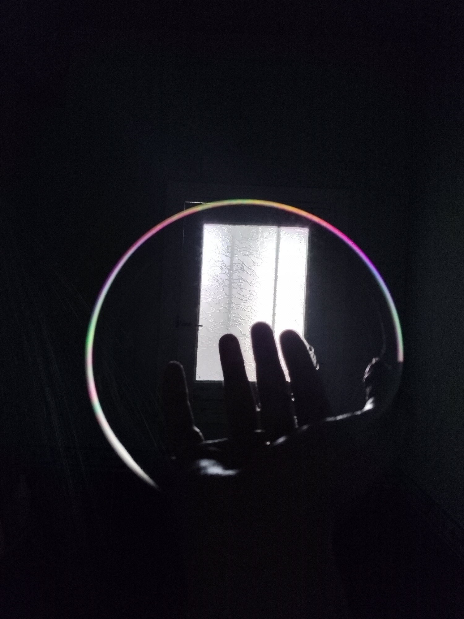 A hand is holding a bubble in front of a window in a dark room. The bubble reflects the light from the window and displays iridescent colors. The window appears to be a paneled door with frosted glass. The overall scene is dark, with the bubble and window being the primary sources of light.