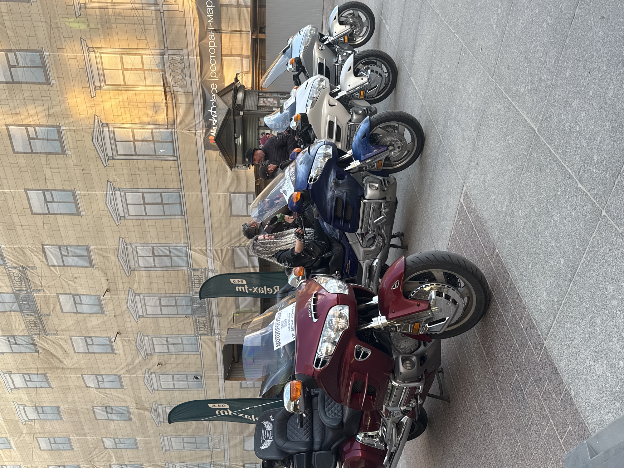 Four touring motorcycles are parked in a row on a paved city square in Saint Petersburg, Russia. The motorcycles are positioned from left to right: a maroon Honda Gold Wing, a dark blue Honda Gold Wing, a white Honda Gold Wing, and a silver Honda Gold Wing. The maroon motorcycle is in the foreground, and its front wheel, handlebars, and part of the fairing are visible. The other three motorcycles are behind it, slightly to the right.
Three people are visible on or near the motorcycles. A woman with long, blonde, braided hair is seated on the dark blue motorcycle. A man wearing a black hat and jacket is standing to the right of the white motorcycle. Another person is partially visible behind the maroon motorcycle, seated on it.
In the background, a large banner depicts the facade of a building with windows and balconies. A smaller banner with Cyrillic text is visible to the left of the maroon motorcycle, and another similar banner is to the right of the dark blue motorcycle. A kiosk with signage that reads "Marketplace | Ресторан-Мар" is positioned behind the motorcycles. The ground is paved with square and rectangular stones. The overall scene appears to be an outdoor event or gathering related to motorcycles.