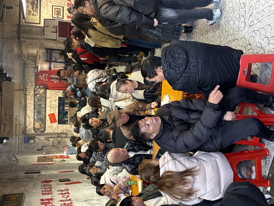 A crowded indoor scene captures a gathering of many people seated closely together at tables. Most are wearing winter clothing, and their attention seems focused on something just out of frame. Red stools provide seating at the tables, and the room has a rustic appearance with aged walls and decorative writings. Some individuals are actively engaged in conversation, while others appear relaxed and contemplative. The arrangement of the people and tables creates a dense and lively atmosphere.