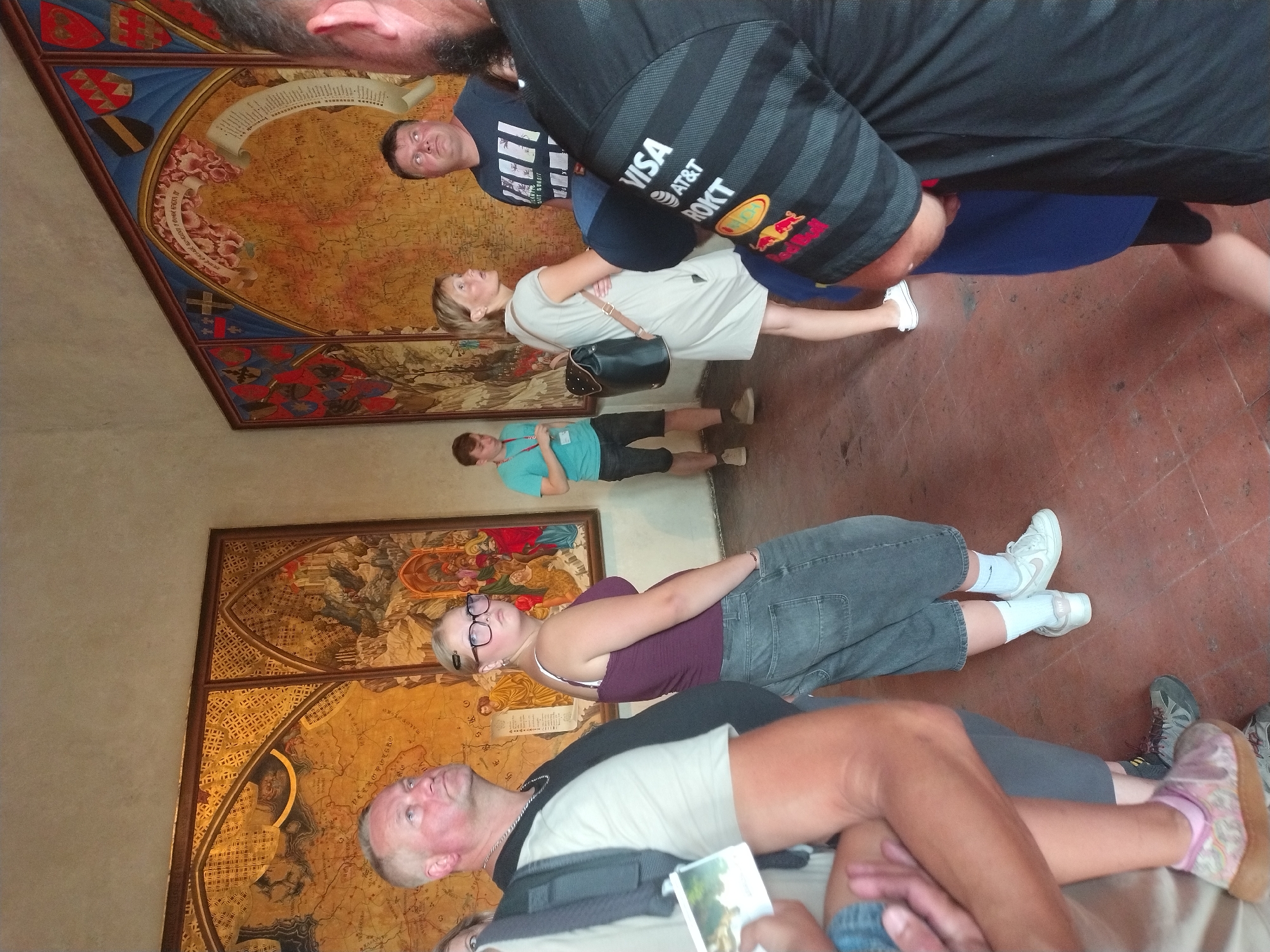 A group of tourists are observing a large, elaborate map displayed on a wall inside a building. The map is highly detailed, featuring cartographic elements, historical scenes, and heraldic symbols. The tourists are diverse in age and attire, with several individuals looking at the map with interest. Some are standing, while others appear to be mid-conversation or movement. The environment seems to be a museum or historical site, indicated by the artwork and the presence of visitors. The lighting suggests an indoor setting, possibly with natural light filtering in from unseen sources.