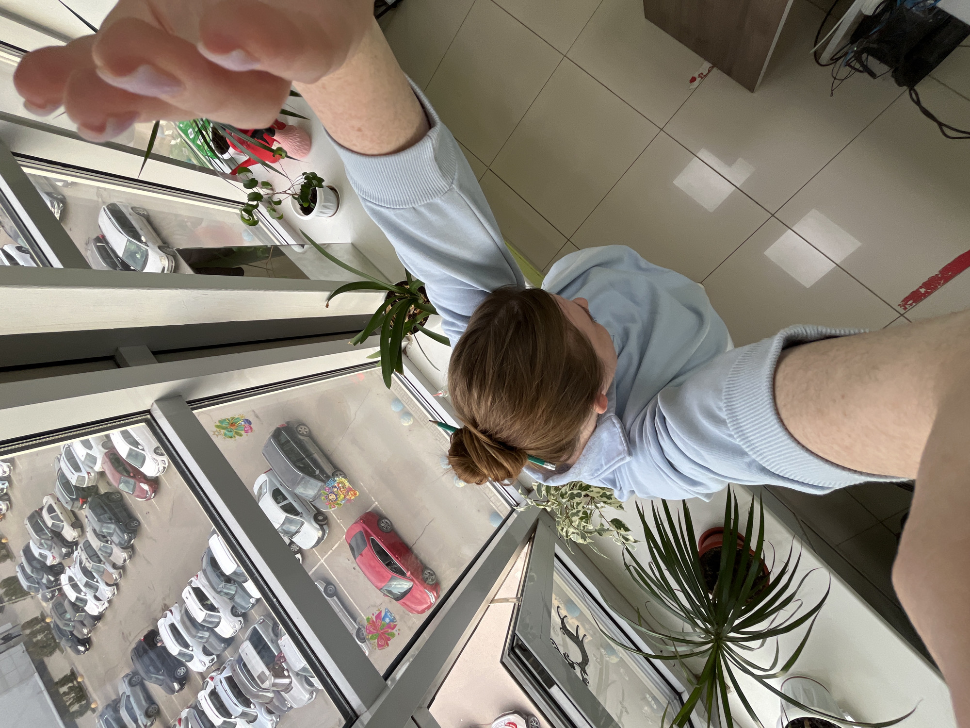 A person wearing a light blue hoodie stands inside a building in what appears to be Klin, Russia, based on the view from the window. The view shows a parking lot filled with various cars, many of which are white. The person is taking a selfie, partially obscuring the scene with their arm and hand. There are several plants placed on the window sills. The floor is tiled and there is a desk in the background.