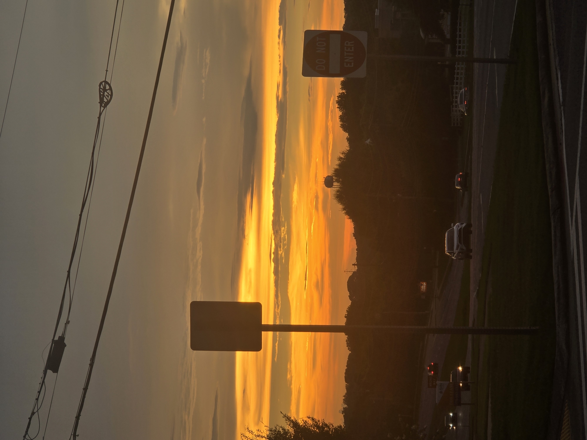 An evening view of a street in Woodstock, United States of America. The sky is a vibrant mix of orange and yellow due to the setting sun, with scattered clouds. In the foreground, a street sign with a blank rectangular plate on a pole is visible, alongside a "DO NOT ENTER" sign. The street is visible with traffic moving, including a white SUV and other cars. Power lines stretch across the top of the frame. In the background, a water tower stands on a hill, surrounded by trees. The overall scene depicts a typical suburban or semi-urban environment at dusk.