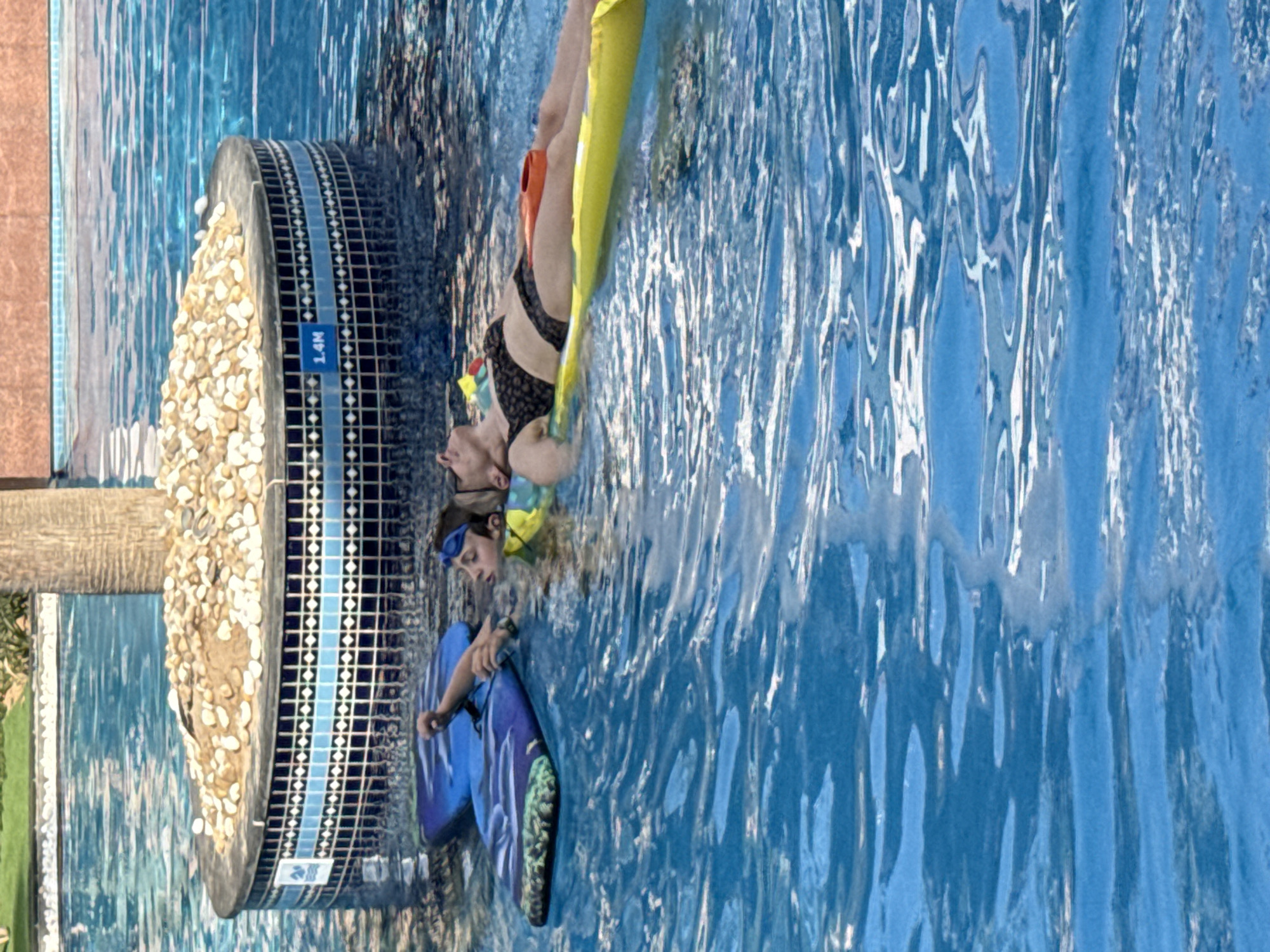 In Dubai, United Arab Emirates, two individuals are seen relaxing in a swimming pool. A young person, wearing blue goggles, is positioned on a blue float with a patterned design, holding onto a portion of a yellow inflatable mattress. Next to them, an adult reclines on the yellow inflatable mattress, wearing a black bikini and with their eyes closed. They are in a large blue swimming pool, with a circular island structure in the background, covered in sand and light-colored pebbles, and tiled with blue and white patterns. A sign on the side of the island structure indicates "1.4M". The water is clear and reflects sunlight, creating a shimmering effect.