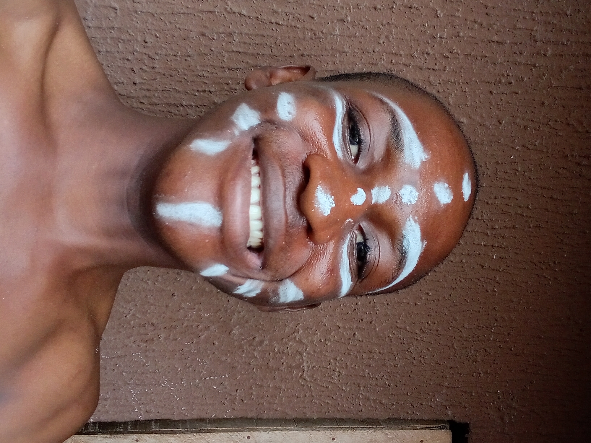 A young person, likely male, with dark skin, is pictured from the chest up, smiling broadly at the camera. The person's head is shaved, and their face is adorned with white face paint. The paint is applied in several distinct patterns: dots on the forehead, a line above each eyebrow extending towards the hairline, cheekbones highlighted with white markings, and vertical streaks on the cheeks. The background is a textured, brown wall, typical of many buildings in Nkpor, Nigeria. A vertical element, possibly a doorframe or window trim, is visible on the right side of the image. The lighting appears to be natural, casting slight shadows and highlighting the sheen of the face paint on the person's skin.
