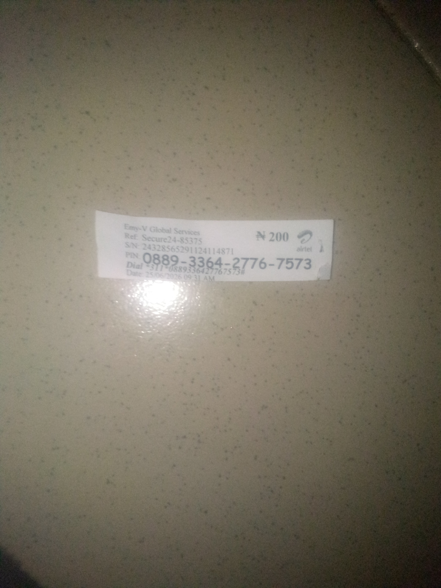 This media file is a close-up, angled photograph of a small white slip of paper with text and numbers on it, placed on a textured, mottled surface. The slip of paper appears to be a recharge card or voucher. The background surface is a neutral, light brown or beige color with many small, dark speckles, possibly indicating a countertop or wall. A bright, diffused light source is creating a significant glare and reflection on the surface, particularly on the lower left side of the slip.
The visible text on the slip includes:
- "Emy-V Global Services"
- "Ref: Secure24-85375"
- "S/N: 24328565291124114871"
- "N 200" (indicating a value of 200 Nigerian Naira)
- The "airtel" logo and word, suggesting it's an Airtel service recharge.
- A prominent PIN number: "0889-3364-2776-7573"
- A dial instruction: "Dial *311*0889336427767573#"
- A date and time: "Date: 25/06/2026 09:31 AM"
The image captures a moment of transaction or record-keeping, likely related to a mobile phone service recharge. The lighting suggests it was taken indoors, possibly with flash. The context of Jalingo, Nigeria, is provided, placing the scene in that specific region. There are no people or animals visible in the image. The focus is entirely on the informational slip.