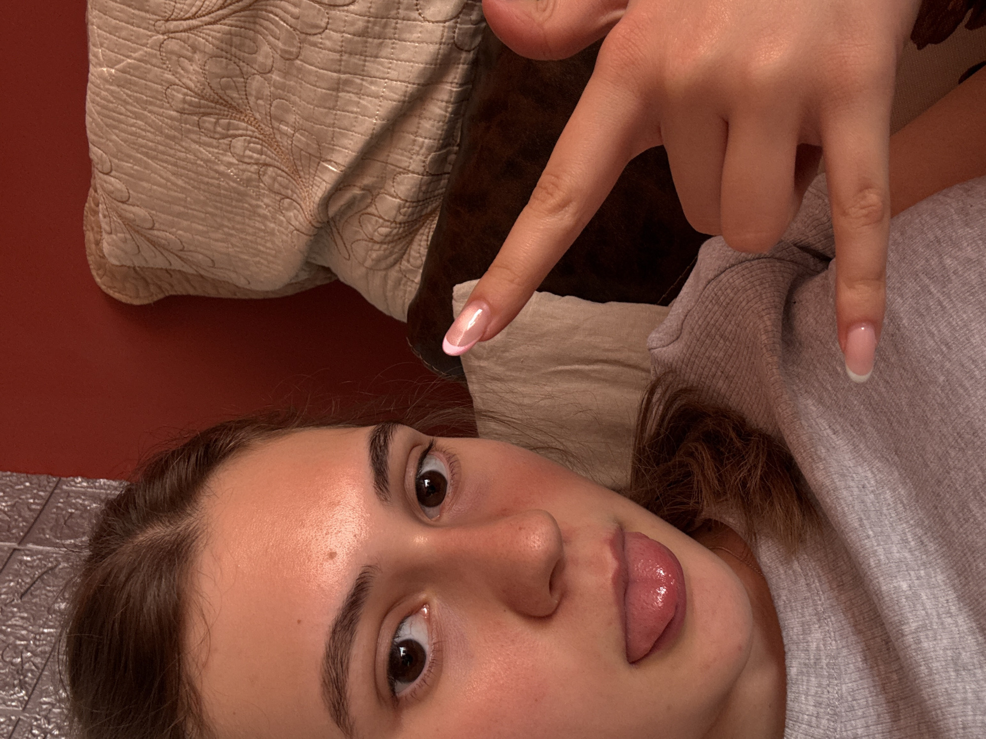 A close-up, eye-level shot features a young woman indoors. She is lying down, oriented with her head towards the bottom of the frame. Her brown hair is partially tied back. Her face is illuminated, highlighting her features: brown eyes, eyebrows, nose, and tongue sticking out playfully. The woman's right hand is raised, fingers extended in a "rock on" hand gesture. Her nails appear to be manicured with a gradient or French tip design.
Behind her, a beige patterned pillow rests on what appears to be a brown cushion or headboard. The background shows a glimpse of a red wall and some tiling.
There is no visual information in the provided image to confirm the location as Panagyurishte, Bulgaria.