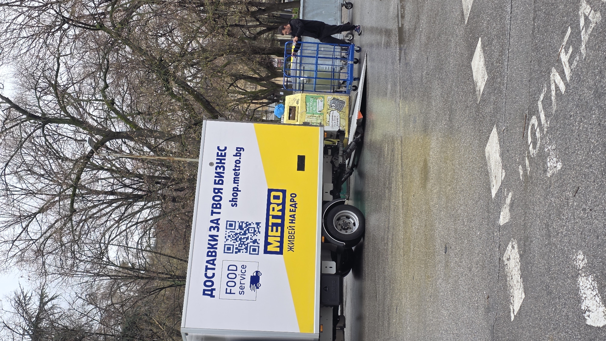 The image depicts a street scene on a wet paved road in Blagoevgrad, Bulgaria. A Metro Food Service delivery truck is parked on the right side of the road, next to pedestrian crossing markings on the street. A man, dressed in dark clothing, is pushing a blue metal cart towards the back of the truck. The truck's advertisement includes the text "ДОСТАВКИ ЗА ТВОЯ БИЗНЕС shop.metro.bg," a QR code, and "METRO ЖИВЕЙ НА ЕДРО." Bare trees are visible in the background. The scene appears to be in a residential or commercial area.