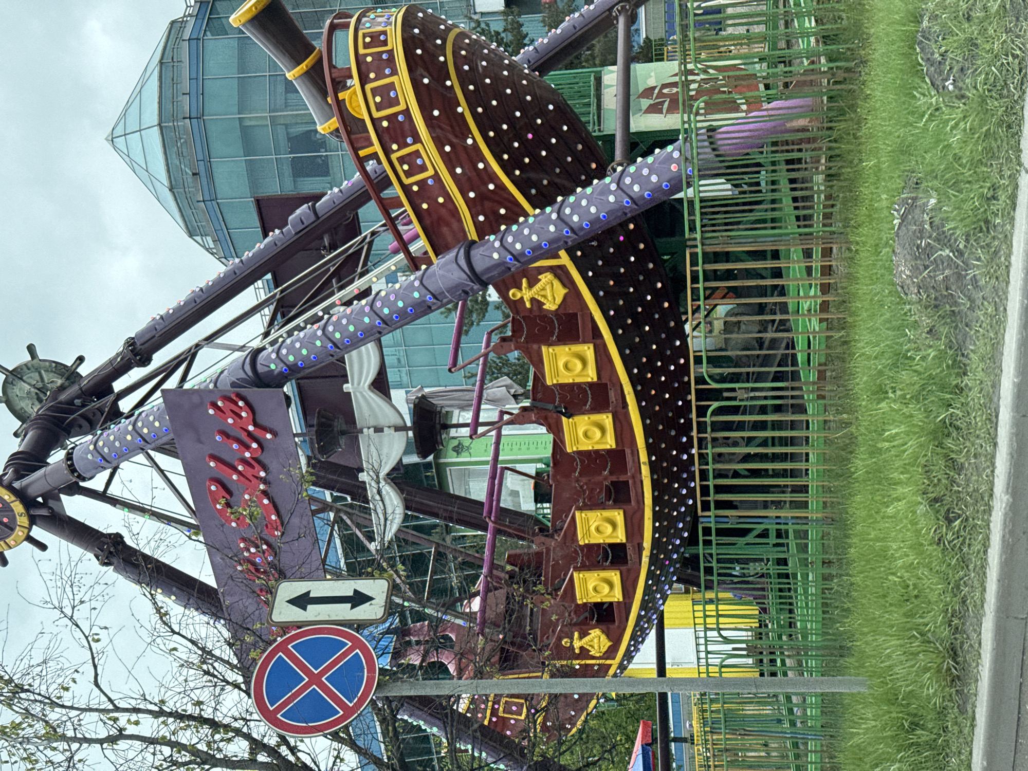 The image shows a carnival ride resembling a pirate ship in Saint Petersburg, Russia. The ride is suspended in the air, with a brown ship-shaped carriage decorated with yellow trim and white lights. Purple metal supports are adorned with strings of colorful lights. A sign with the ride's name in red script is visible. Below the ride, there's a street sign with arrows pointing in opposite directions and a "no parking" sign. A green metal fence separates the ride from a grassy area and a sidewalk. In the background, there's a modern glass building and some trees. The sky is overcast.