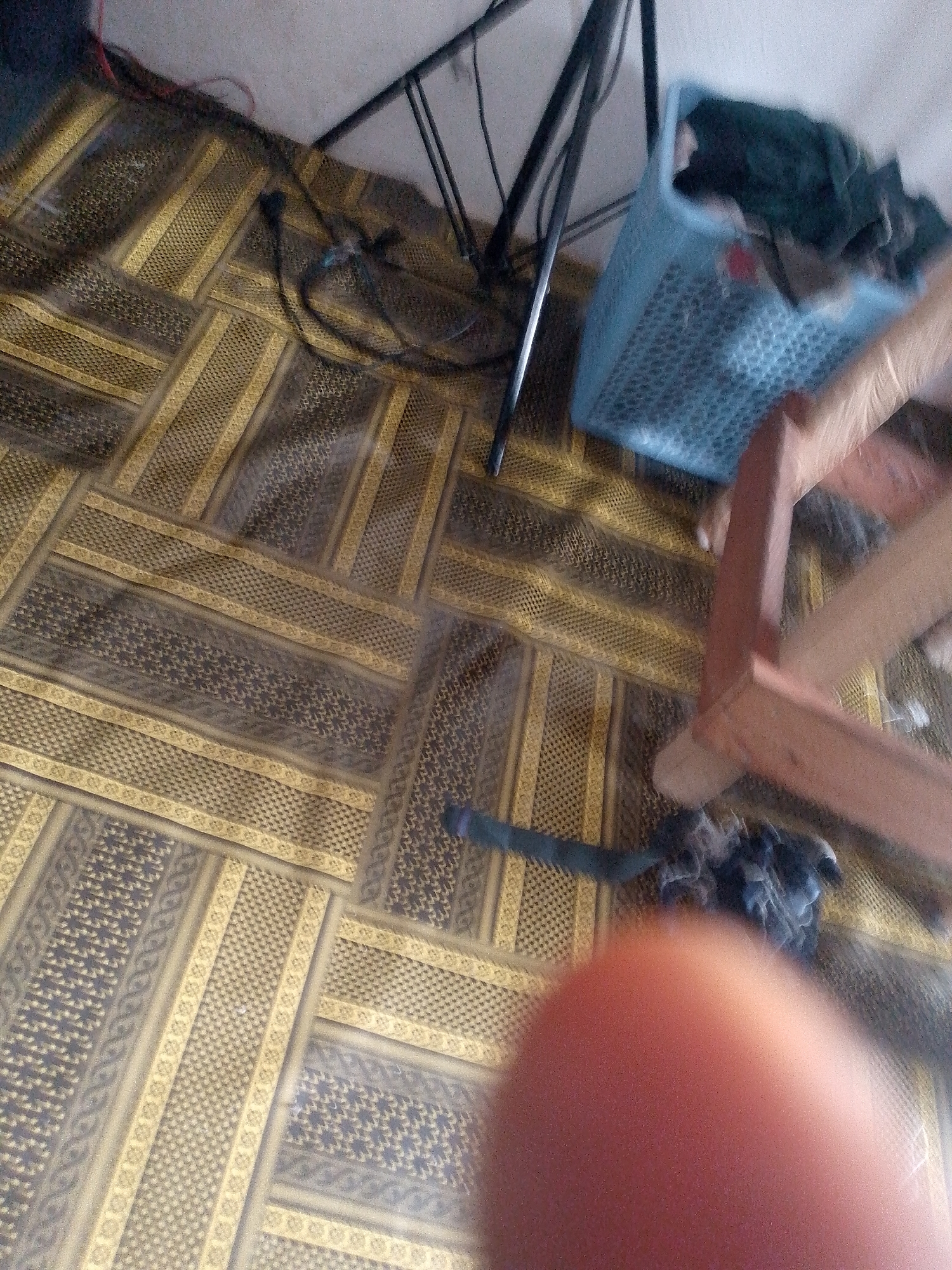 The image shows a floor with a patterned carpet and a blue plastic basket containing dark-colored fabric. A wooden structure with reddish-brown legs is partially visible on the right. A black cable is draped on the carpet. The scene is dimly lit. The location is identified as Jauro Yinu, Nigeria.