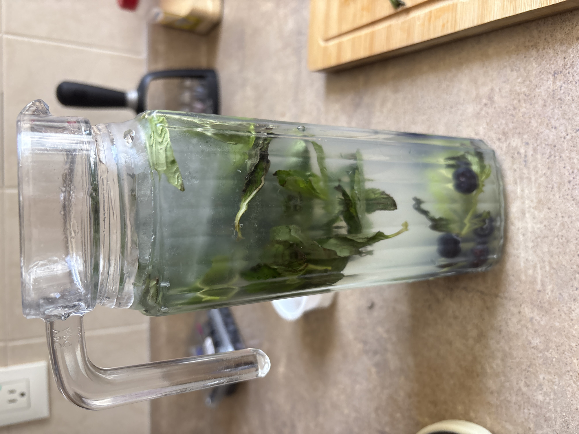A glass pitcher filled with water, basil leaves, and blueberries sits on a countertop. In the background, there is a wooden cutting board, a grater, and an electrical outlet. The location is Huixquilucan de Degollado, Mexico.