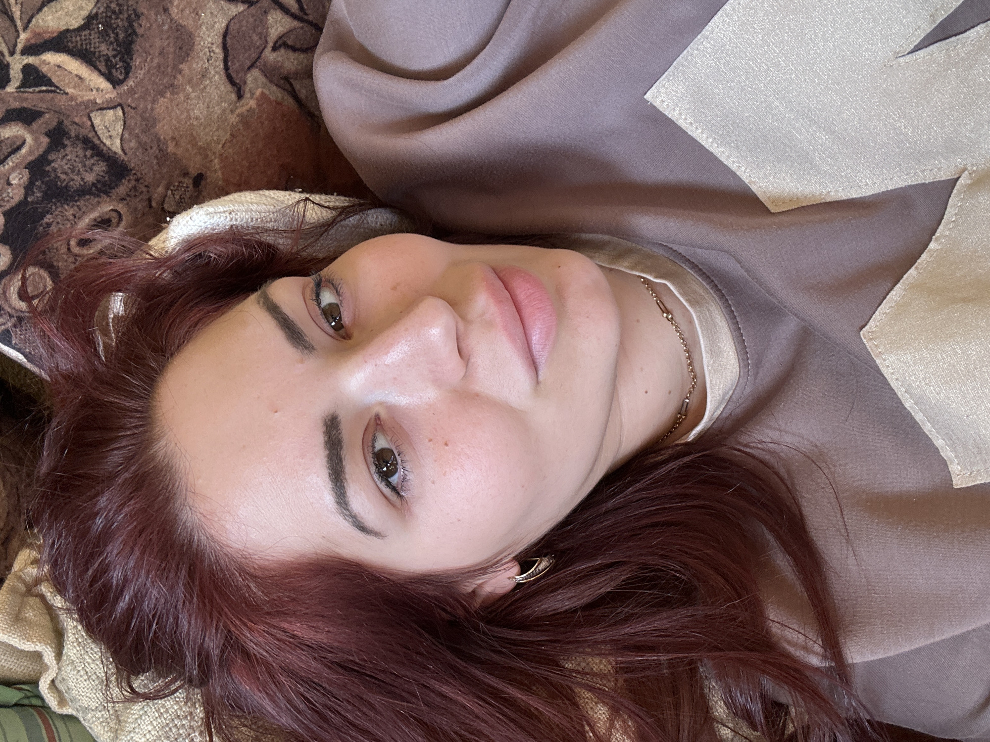 A woman with auburn hair is lying on a patterned surface. She is wearing a tan top with a light beige trim. Her face is centered in the frame, showing her eyes, nose, and lips clearly. She has fair skin and visible freckles. A gold chain necklace is visible around her neck, and she is wearing gold hoop earrings. The location is not discernible from the image.