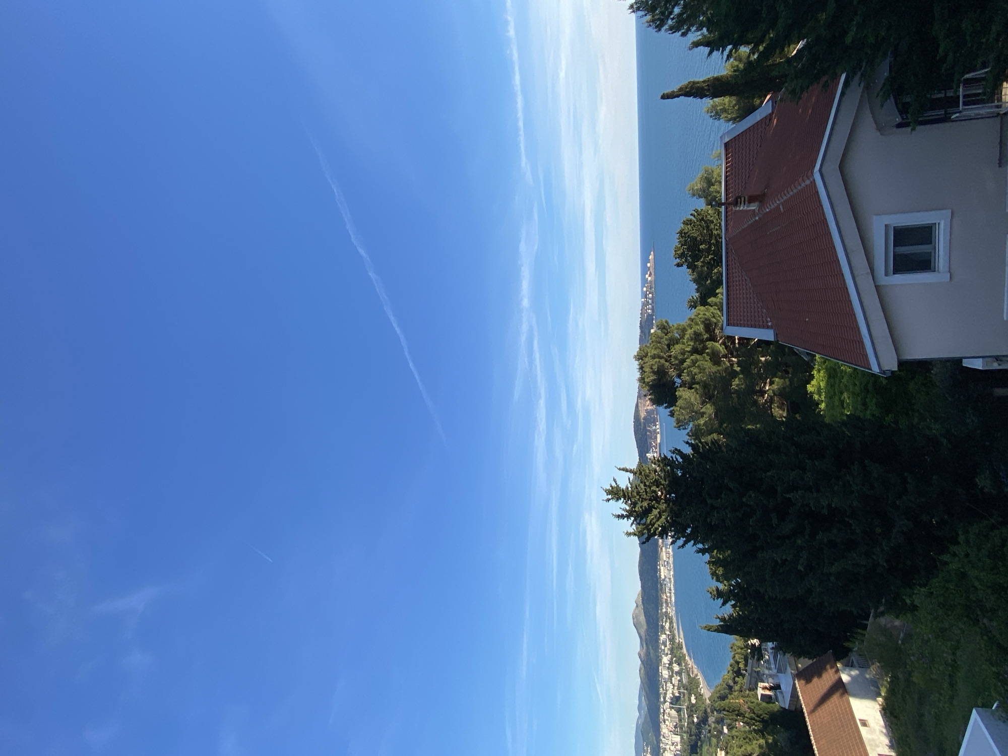 The image shows a coastal view in Šušanj, Montenegro. The sky is blue with contrails. The Adriatic Sea is visible in the distance, with a peninsula and urban development along the coast. A building with a red tile roof is on the right side of the image, partially obscured by trees. The foreground includes foliage.