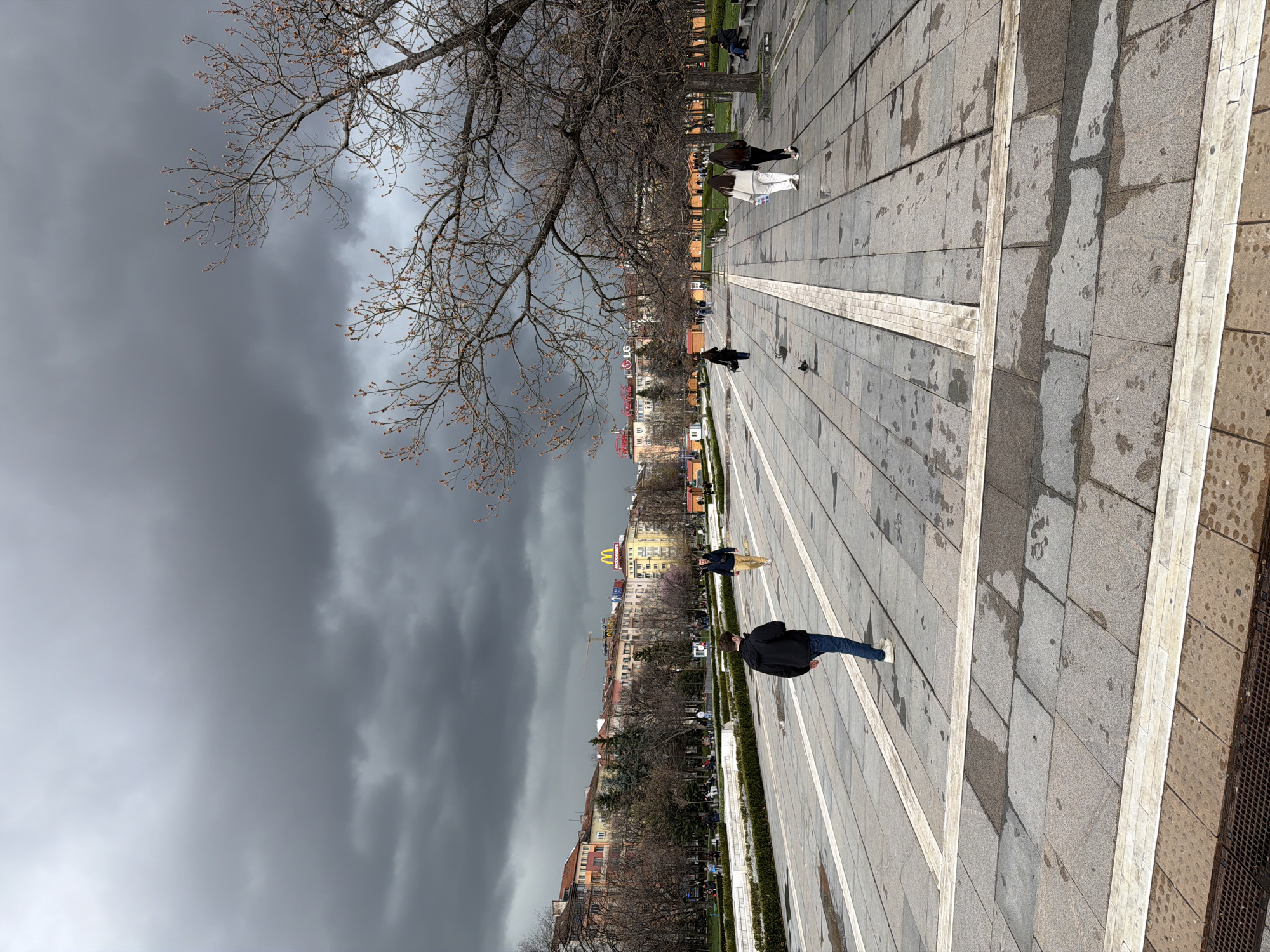 The image shows a wide pedestrian area in Sofia, Bulgaria, likely near the National Palace of Culture. The ground is paved with grey stone tiles, punctuated by long strips of lighter-colored stone. The sky is overcast with dark, stormy clouds. Several people are visible walking along the promenade, including individuals in dark jackets and jeans. There are trees lining the walkway, some bare-branched. In the background, buildings are visible, and one building bears the golden arches of a McDonald's restaurant. The ground appears wet, suggesting recent rain.