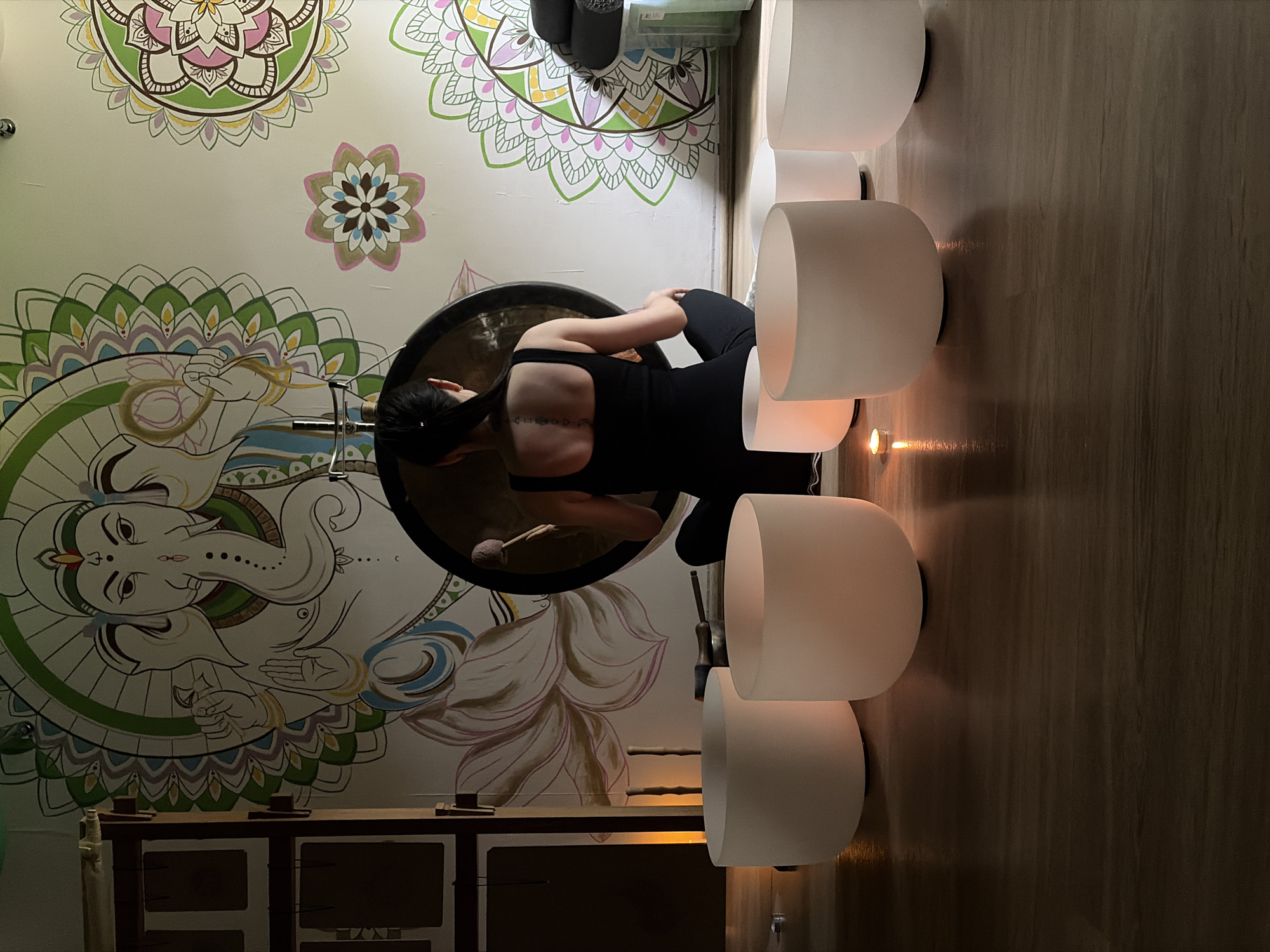 An indoor scene in Chaikovsky, Russia, depicts an individual engaged in sound healing or meditation. A female figure, viewed from the back, is seated cross-legged on a light-colored floor, wearing a black tank top and dark pants. Her dark hair is tied back, revealing a vertical tattoo on her upper back. She is positioned directly behind a large, dark circular gong suspended on a stand, holding a mallet with a light-colored, fuzzy head, poised as if to strike the instrument.
In the foreground, arranged on the wooden floor, are six frosted white crystal singing bowls of various sizes, illuminated from within or by ambient light, casting a soft glow. A small lit tea light candle rests on the floor among the bowls, contributing to the ambient lighting. Additional mallets or tools are visible on the floor behind the bowls.
The white wall in the background is decorated with a large, colorful mural centered on the Hindu deity Ganesha, depicted with a trunk, multiple arms, and a halo-like element behind his head. The mural incorporates elements like lotus flowers and intricate mandala patterns in shades of green, blue, pink, yellow, and brown. To the far left, a dark wooden structure with multiple compartments is partially visible. On the right, stacked containers with rolled mats or towels are present. The overall lighting is dim and atmospheric.