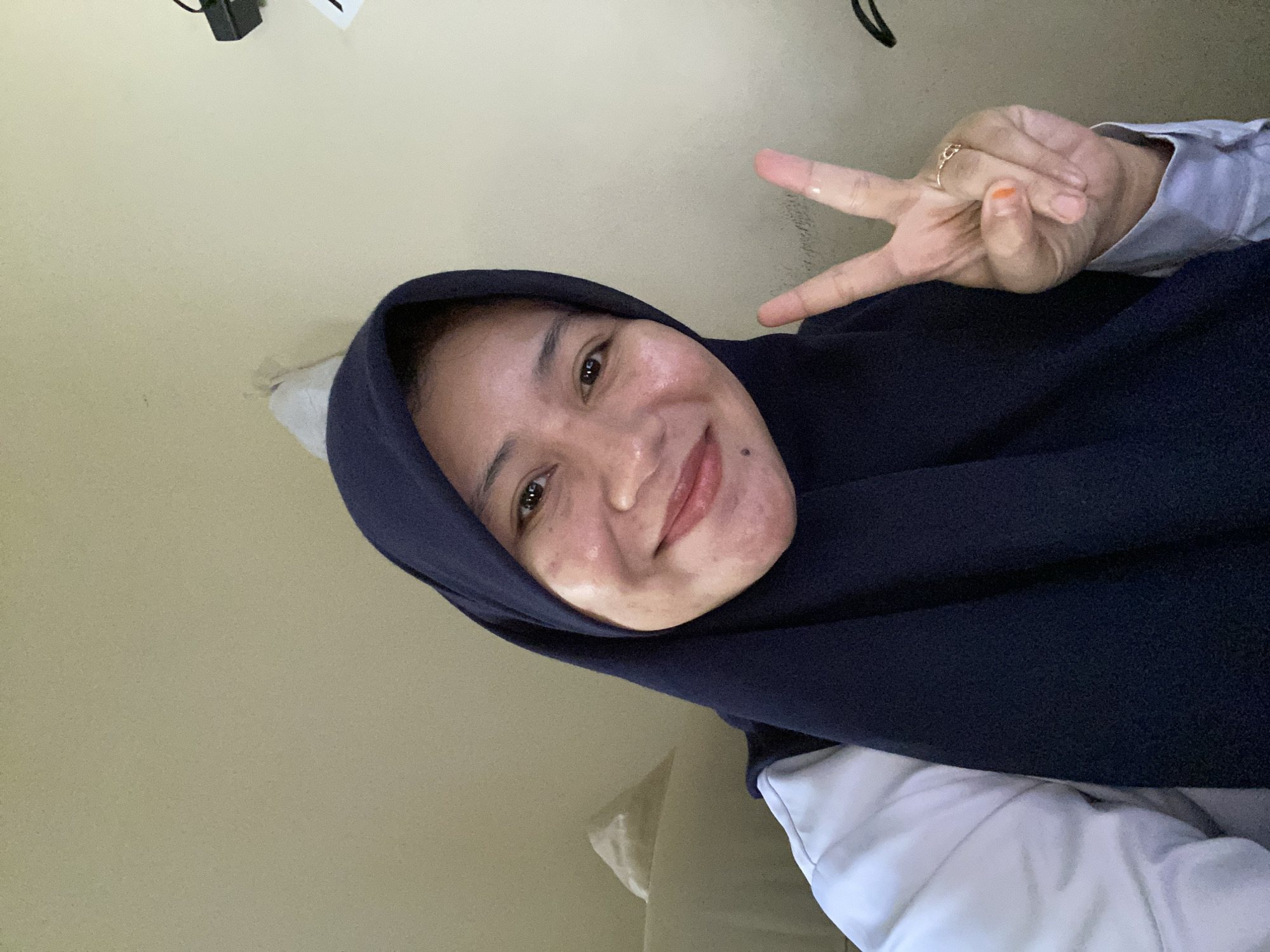 A medium shot selfie shows a woman wearing a navy blue hijab and a white collared shirt. She is smiling and holding up a peace sign with her right hand. The background is a light beige wall, with some indeterminate objects visible in the upper right corner. The image appears to have been taken indoors, likely in a home or office setting. There is no visible indication of the location being in Banjarbaru, Indonesia.