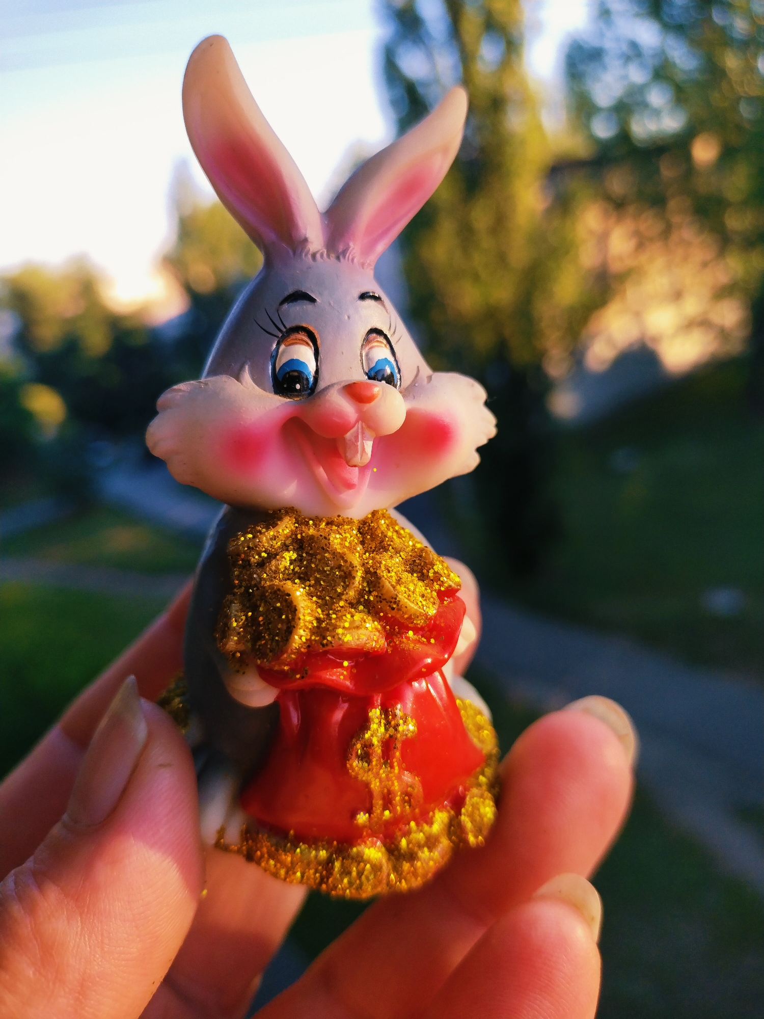 A close-up, outdoor, eye-level shot shows a person's hand holding a small, anthropomorphic rabbit figurine. The figurine has a grey body, large pink-lined ears, wide blue eyes with black eyelashes, rosy cheeks, and an open mouth revealing two front teeth. It is holding a red bag adorned with gold glitter and a dollar sign, filled with what appears to be gold coins or glitter. The background is blurred, suggesting a shallow depth of field, and depicts trees and sky, indicating an outdoor setting. The overall lighting suggests it is daytime, possibly late afternoon due to the warm tones. The image was captured in Horishni Plavni, Ukraine.