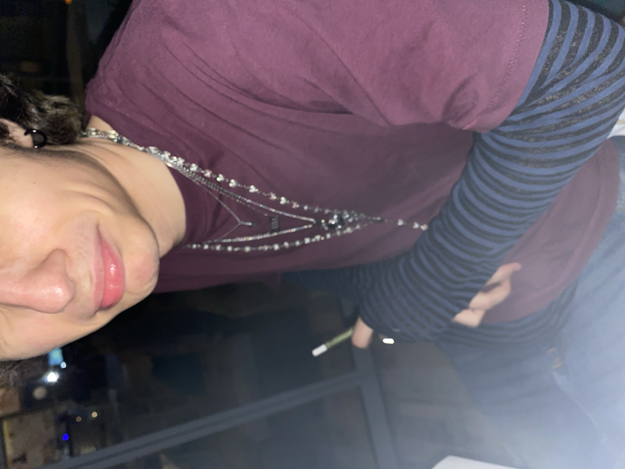 A person is facing toward the left of the frame, wearing a maroon shirt, a striped blue and black undershirt, and a silver necklace. The person's right hand is visible holding a cigarette or small white stick. Through a window, blurred lights of a city at night are visible, with horizontal bars of an unseen balcony or window frame running behind. It is impossible to identify the location in Stara Zagora, Bulgaria.