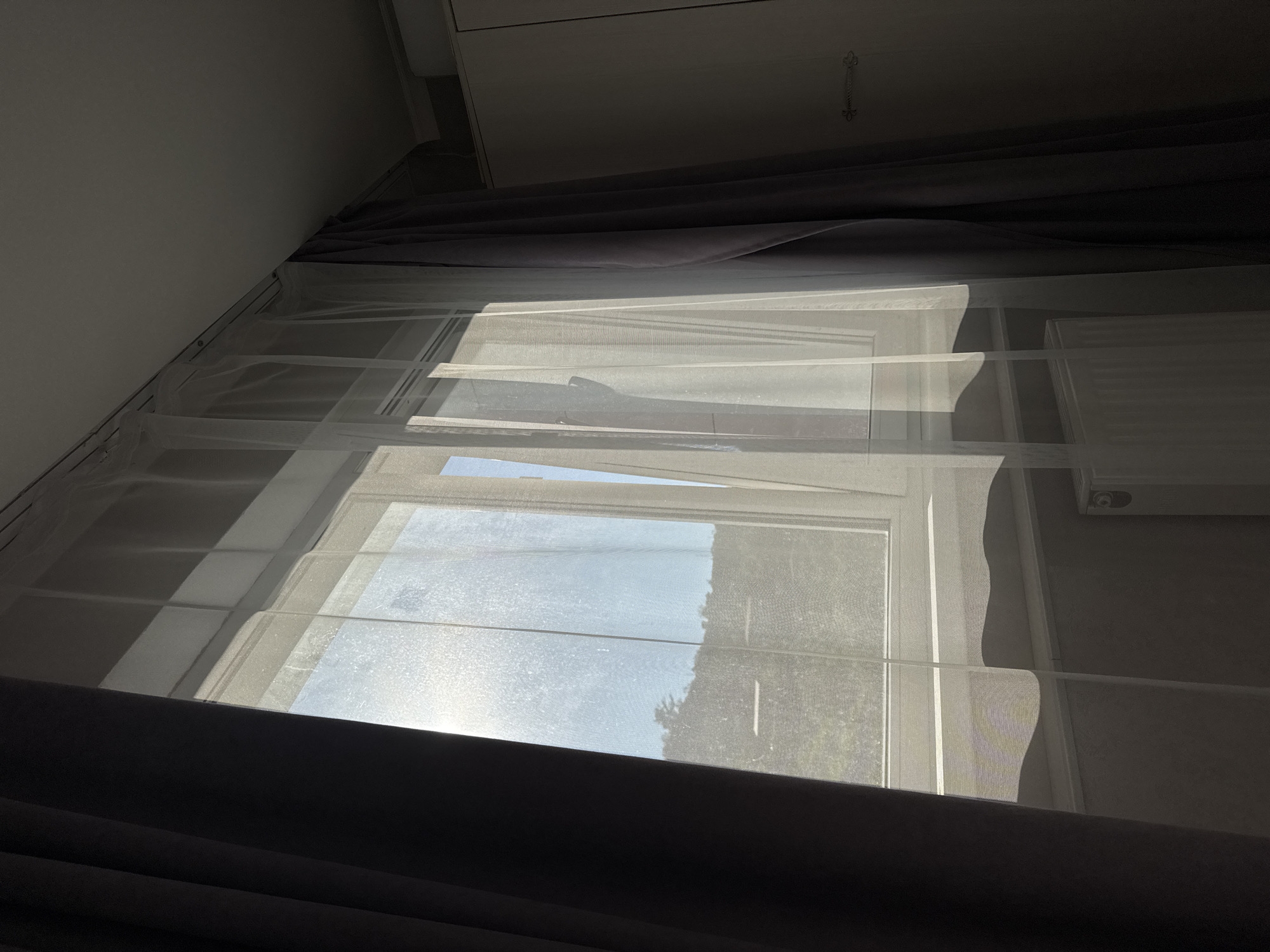 The image displays an interior room in the city of Sochi, Russia, featuring a window as the primary light source. A multi-pane window with a white frame is positioned centrally. It is dressed with two layers of curtains: an inner sheer white fabric curtain and an outer, heavier dark grey or purplish opaque curtain. The outer curtains are pulled back, exposing the window. Intense sunlight streams through the window, diffused by the sheer fabric, creating significant glare, particularly on the left side. Through the translucent curtains, an outdoor environment is visible, characterized by green foliage and trees under a clear, light blue sky. Faint horizontal structures are discernible in the distant background. A white windowsill is present below the window, and a portion of a white radiator is visible in the lower left section. The room's ceiling, light in color, is visible at the top, equipped with a curtain track. On the far right side of the image, a light-colored wardrobe or cabinet with a metallic, ornate handle is partially visible, primarily in shadow. The interior walls are a neutral, muted tone, and are also predominantly in shadow. No human subjects, animals, or specific actions are depicted within the scene.