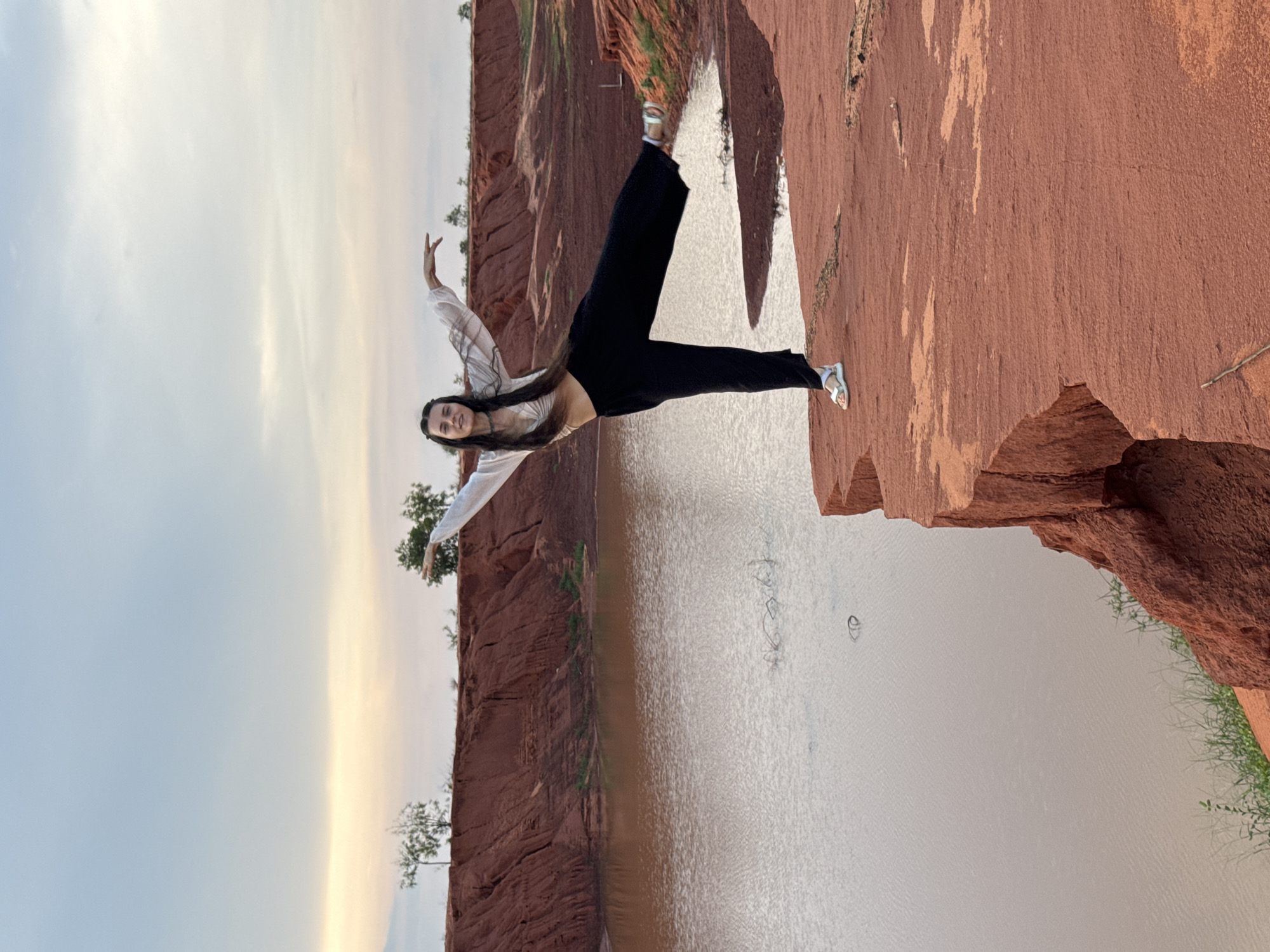 A young woman is captured mid-pose on a reddish-brown cliff edge overlooking a body of water, likely at either sunrise or sunset given the soft, warm light. She stands gracefully, balancing on her left leg with her right leg lifted high and her arms extended outward, giving the impression of flying or dancing. Her long dark hair flows down her back, and she wears a light-colored, possibly sheer, long-sleeved top over a dark crop top, paired with black wide-leg pants and light-colored sandals. A clear smile on her face conveys joy and freedom.
The scene is dominated by dramatic red-orange cliffs, suggesting a distinctive geological formation like those found in Mui Ne, Vietnam. Below the woman, a calm body of water reflects the sky, with ripples indicating gentle movement. On the opposite side of the water, more reddish cliffs with sparse green trees and vegetation are visible. The sky above transitions from a pale blue to soft orange and yellow hues near the horizon, typical of the golden hour. The image evokes a sense of adventure and connection with nature. No text is visible.