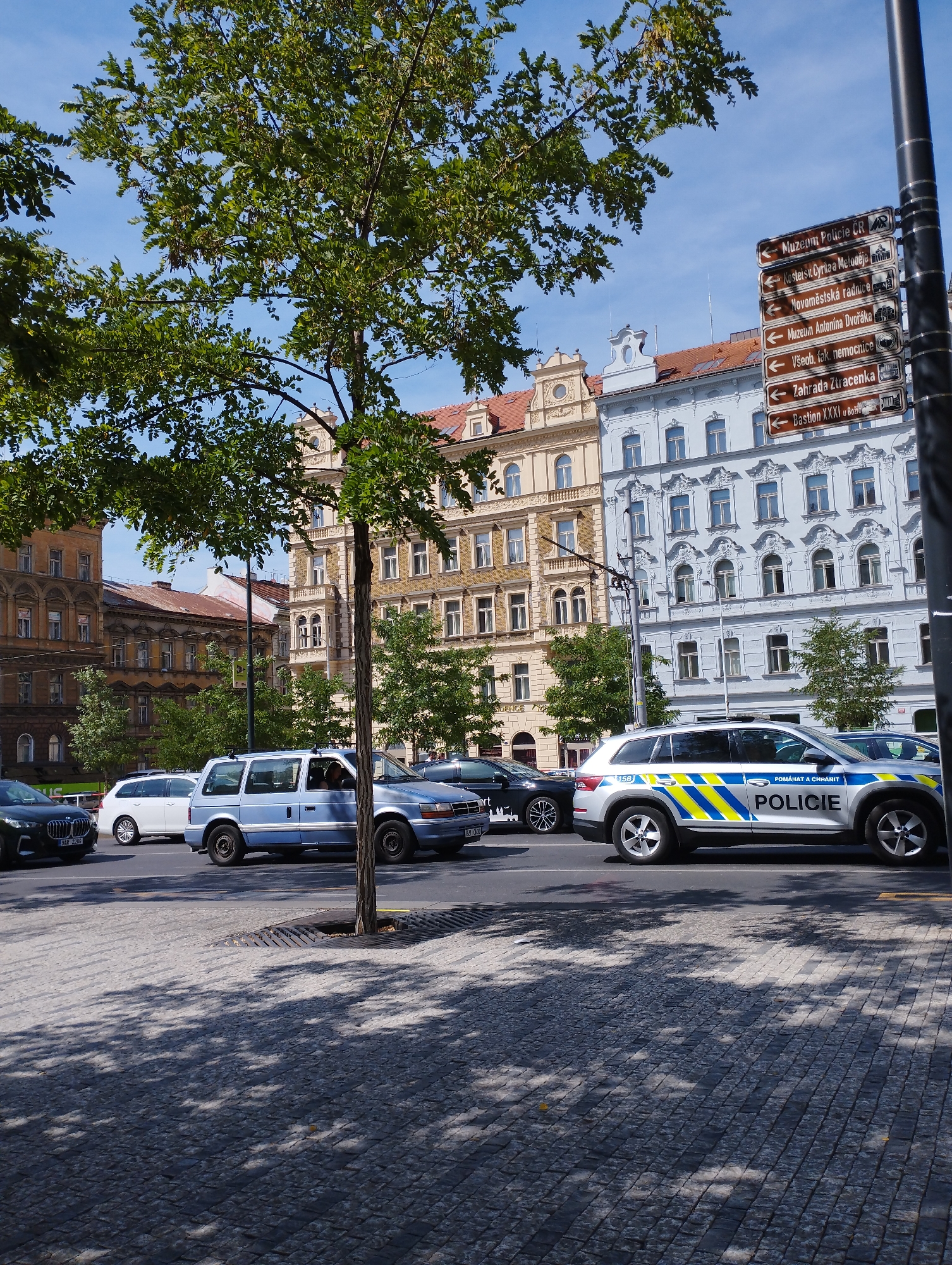 The image depicts a street scene in Prague, Czechia, on a sunny day. In the foreground, a cobblestone sidewalk is partially shaded by the leaves of a deciduous tree on the left. Several cars are visible on the road. A silver police car with "POLICIE" written on its side is prominently displayed, facing right. To its left, a light blue minivan is also parked. Further to the left, a white SUV and a dark sedan are partially visible. In the background, historic buildings with ornate architecture line the street, featuring light yellow and light blue facades. A signpost with multiple directional signs in Czech is affixed to a pole on the right side of the image. The sky is clear and blue.