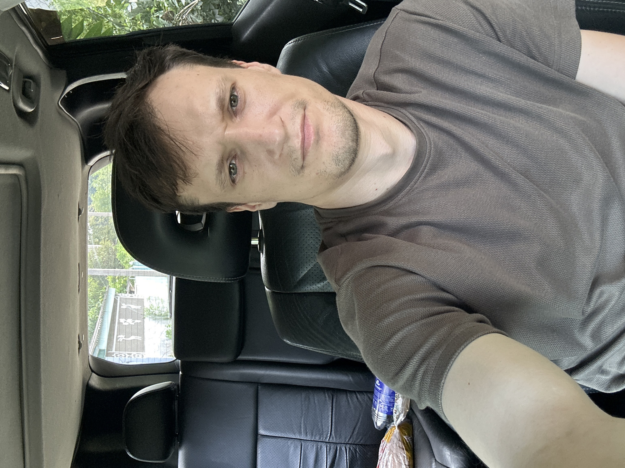 A man with short brown hair and stubble is sitting in the driver's seat of a car. He is wearing a brown t-shirt. The car's interior is visible, with black leather seats and the ceiling of the car. Through the windshield, a building with some figures painted on it is visible. The image was taken in Makiv, Ukraine.