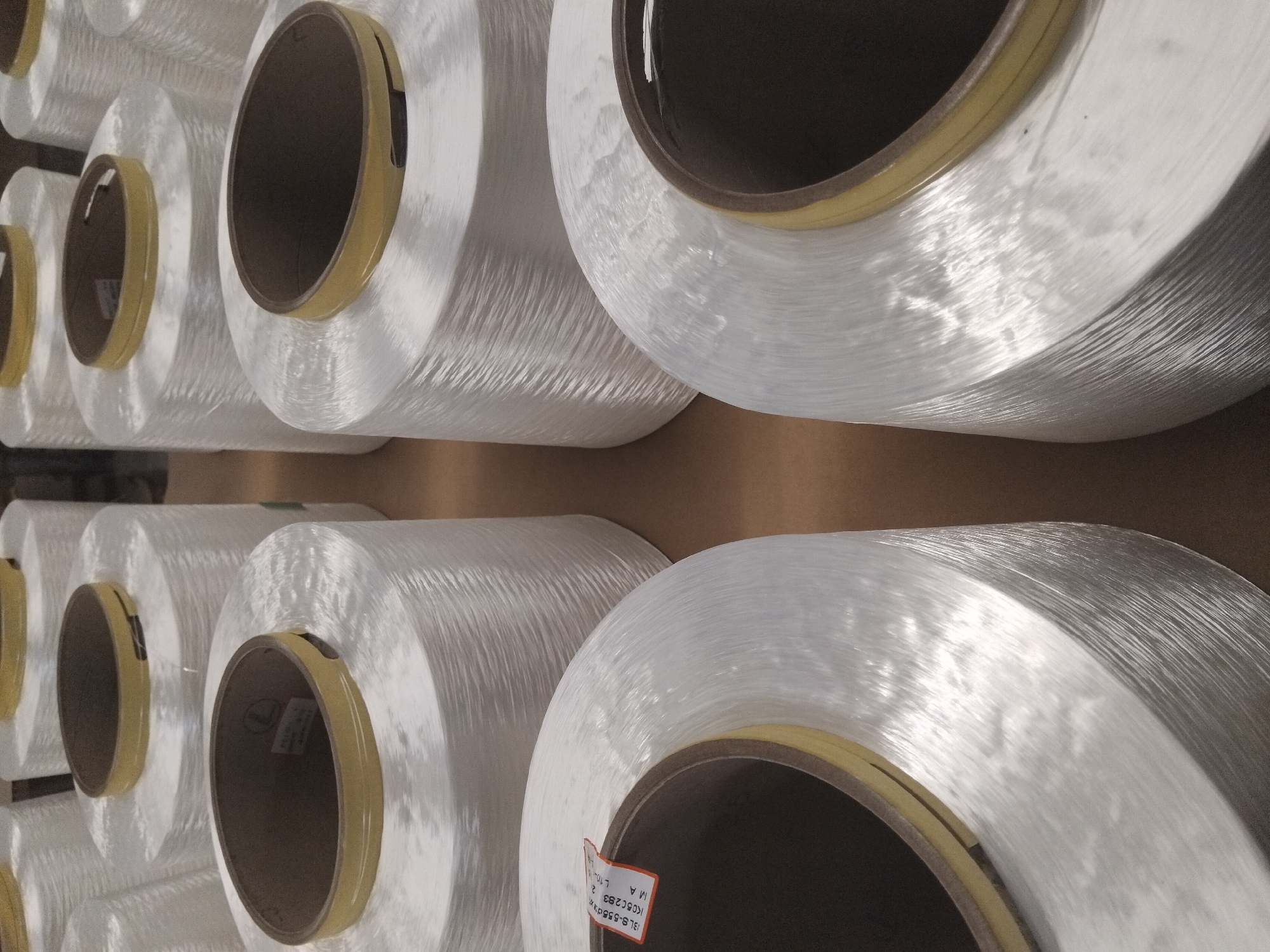 This media file shows several large spools of white, translucent material, likely fiberglass yarn or thread, arranged in rows. The spools are cylindrical with cardboard cores and yellow tape around the edges. The material on the spools is wound tightly, creating a textured, slightly shimmering surface. The lighting suggests an indoor setting, possibly a warehouse or factory, with light coming from above and to the right, creating highlights and shadows that emphasize the texture of the material. The arrangement of the spools indicates a manufacturing or storage environment. Some of the spools have labels attached to their cores, though the text on these labels is mostly illegible. One label is partially visible and appears to have alphanumeric characters and possibly some Dutch text, given the location context of Deurne, Netherlands. The overall impression is one of industrial production and supply. No people are visible in the image, and the scene is static. The time of day and weather are not discernible from the image.