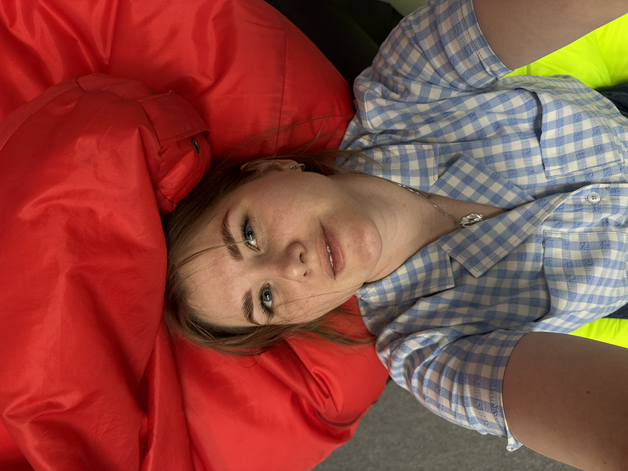 A young adult female is lying on a red padded surface, possibly a beanbag chair or cushion. She is wearing a light blue and white checkered button-down shirt. She is fair-skinned with light brown hair. She is wearing a silver necklace with a pendant. The background includes a green surface and a gray surface. The location is not discernable from the image; the city of Perm, Russia, cannot be confirmed from the visual data.