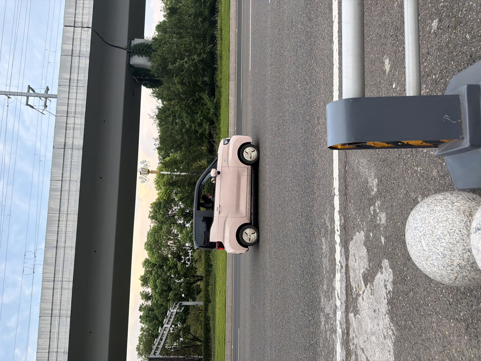 In Jiangjin, China, a pink electric microcar with a black roof is driving on a road under an elevated structure. The car has tinted windows and a driver is visible inside. The road surface is asphalt, with white lane markings. To the left of the road, there is a grassy median strip with trees. The elevated structure appears to be a highway or railway bridge, made of concrete. Power lines are visible above the bridge. In the foreground, a grey guardrail with yellow reflective markers and a spherical stone bollard are present. The sky is overcast with some hints of sunset colors.