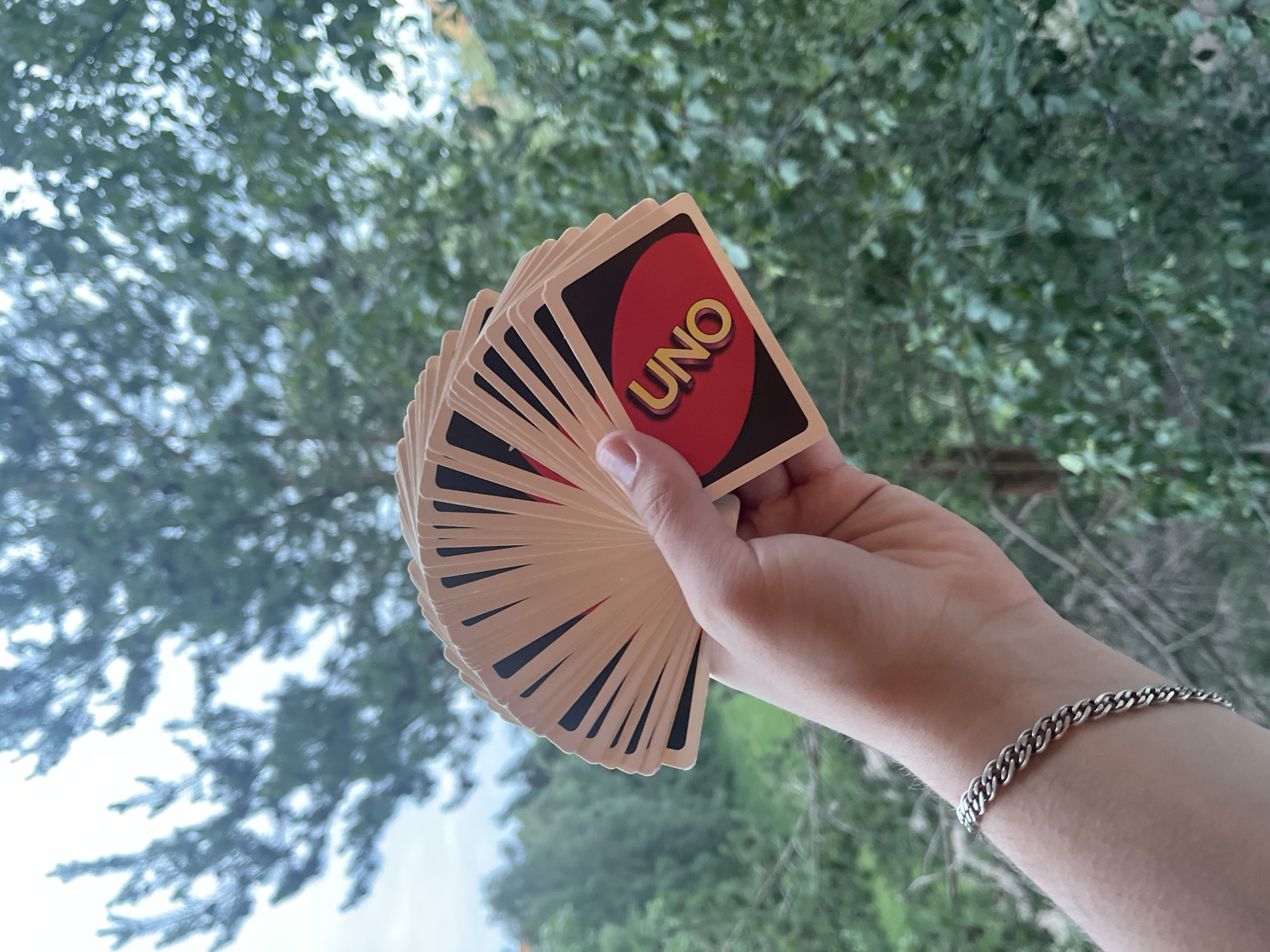 A hand with a silver bracelet holds a fanned deck of Uno cards outdoors. The background is blurred, showing green foliage and a cloudy sky. The Uno logo is visible on one of the cards.