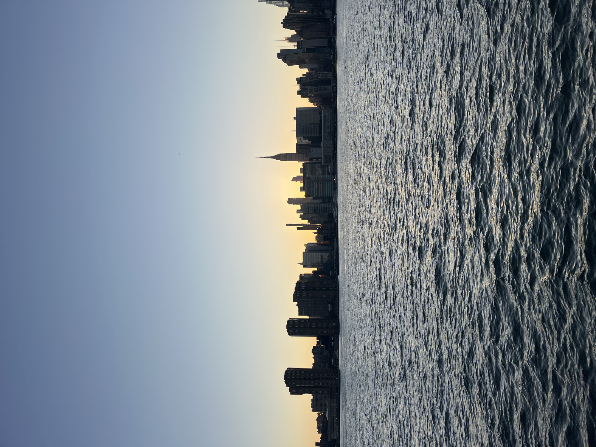 The image depicts a skyline of New York City, specifically Manhattan, viewed across a body of water at sunset. The sky transitions from a clear, pale blue at the top to a warm yellow and orange hue near the horizon where the sun is setting, casting a glow behind the buildings. The water in the foreground is dark and choppy, with reflections of the sky and city lights.
The skyline is dominated by silhouetted skyscrapers, including the iconic Empire State Building, which stands out with its distinct spire. Other buildings of varying heights and architectural styles are visible, forming a dense urban landscape. The buildings appear dark due to the backlight from the sunset. The perspective suggests the photo was taken from across the East River, possibly from Brooklyn or Queens, looking towards Midtown Manhattan. There are no people or specific actions visible in the scene; it is a static, panoramic view of the city at dusk.