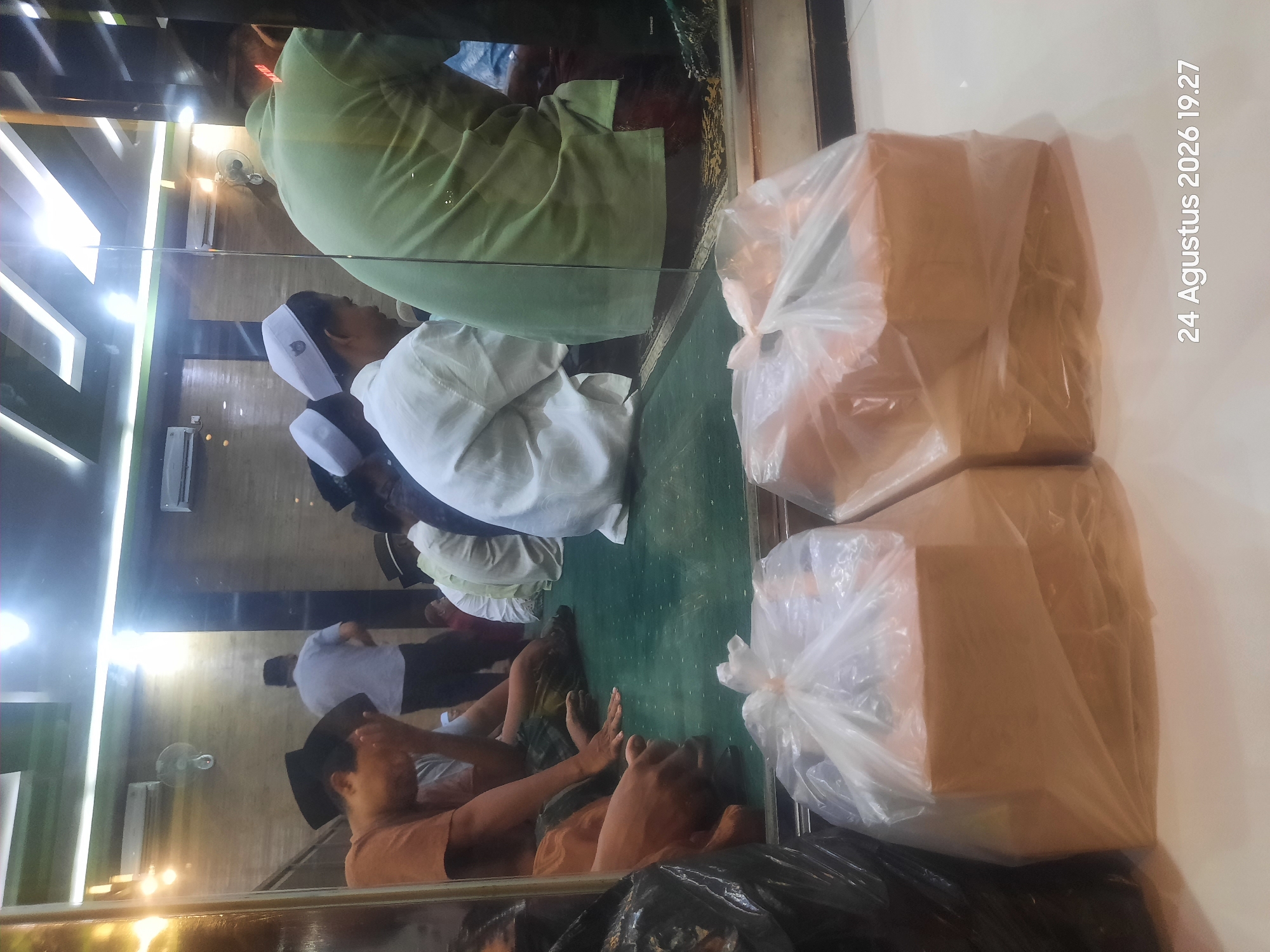 The image depicts an indoor scene, likely a mosque or prayer hall, with several individuals seated on a green carpeted floor. The architecture suggests a modern building with a reflective glass partition in the foreground. In the foreground, two plastic-wrapped packages are prominently displayed. Behind the glass partition, several people are visible, some wearing traditional Islamic attire, including white peci hats and white robes. One person in the background is standing and appears to be walking away. The lighting is bright, with overhead fluorescent lights casting reflections on the glass and ceiling. The overall atmosphere appears to be peaceful and communal. The date stamp at the bottom right corner reads "24 Agustus 2026 19.27", indicating the photograph was taken on August 24, 2026, at 7:27 PM. The location in the city of Sukokerto, Indonesia, is not explicitly visible in the image, but the date format and script suggest an Indonesian context.