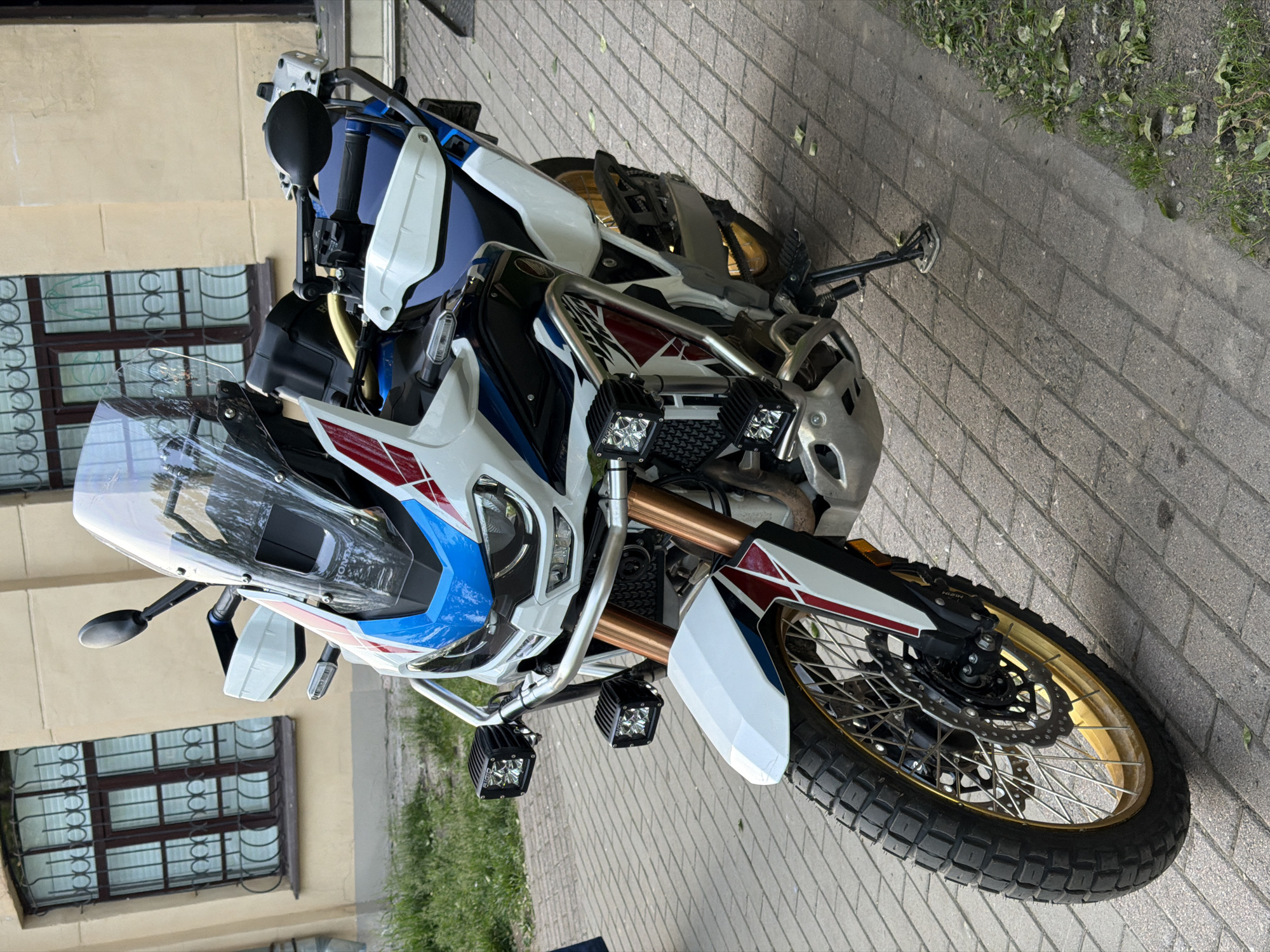 A white, blue, and red Honda Africa Twin Adventure Sports motorcycle, featuring gold-colored spoked wheels and knobby tires, is parked on a grey brick-paved sidewalk. The motorcycle is equipped with a tall clear windscreen, white handguards, silver tubular crash bars, and four square black LED auxiliary lights (two on each side of the crash bars, one bearing "RIGID" branding). Copper-colored front forks are visible. The motorcycle rests on its deployed side stand, angled towards the right foreground.
In the background, a light yellow/cream-colored building with dark brown-framed windows stands. Each window is fitted with decorative, dark-colored wrought iron security grilles exhibiting circular and linear motifs. Reflections of greenery and sky are present in the windowpanes. To the right of the sidewalk, a narrow strip of ground with sparse green grass and scattered dry leaves is visible. No individuals or specific actions are discernible within the frame. The scene is located in the city of Saint Petersburg, Russia.